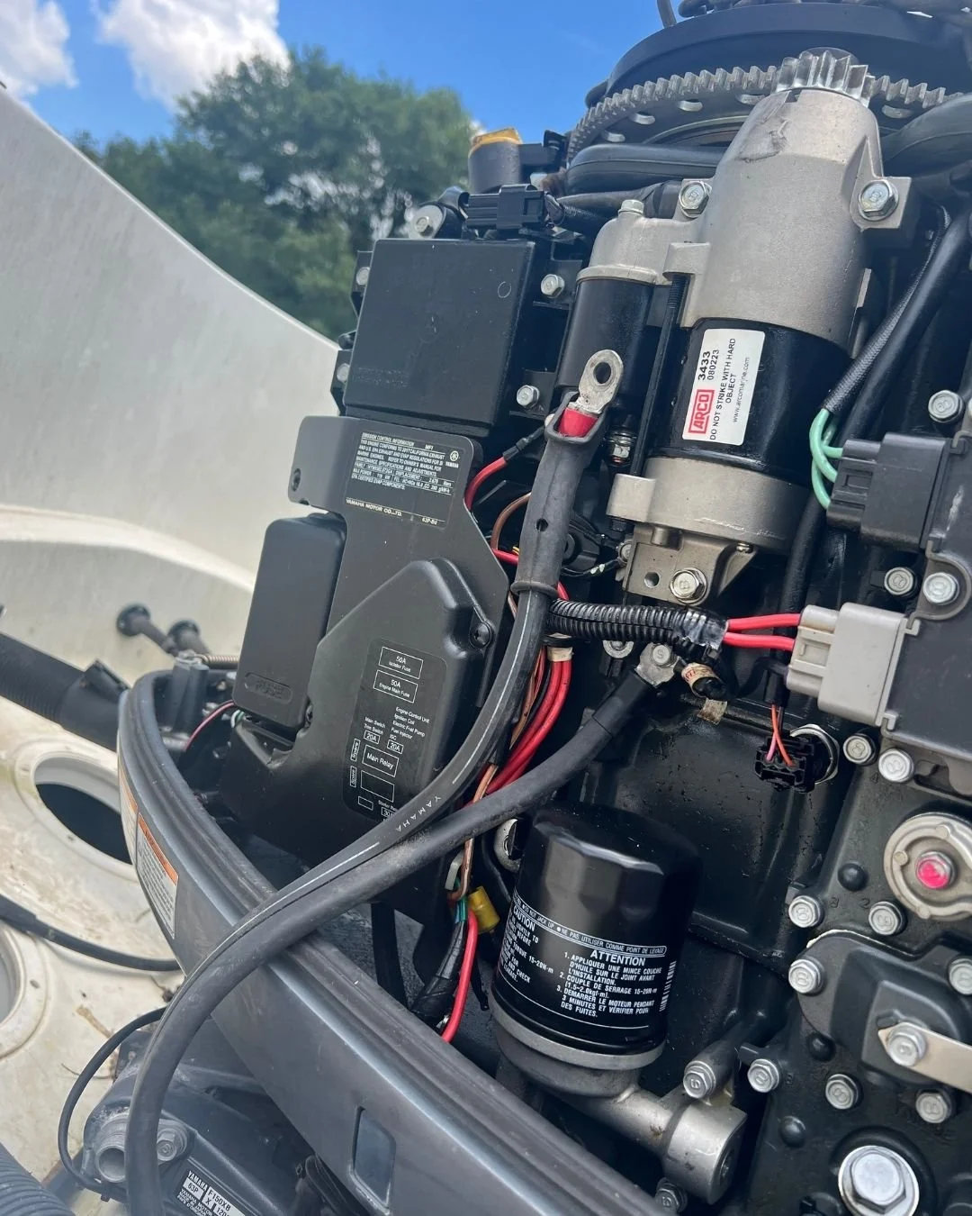 Close-up view of the internal components of a boat engine, including wiring, filters, and electronic parts, with an outdoor background and a partly cloudy sky.