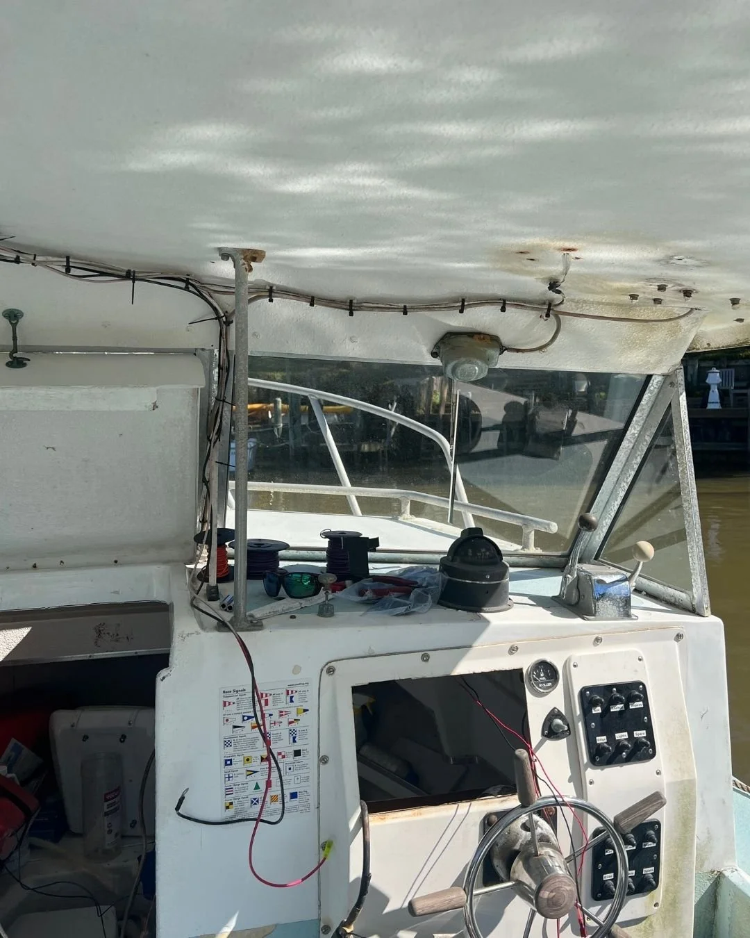 View of a boat dashboard with various wiring, a steering wheel, and navigation controls; part of the boat's interior with a glass windshield and a view of a marina and water outside.