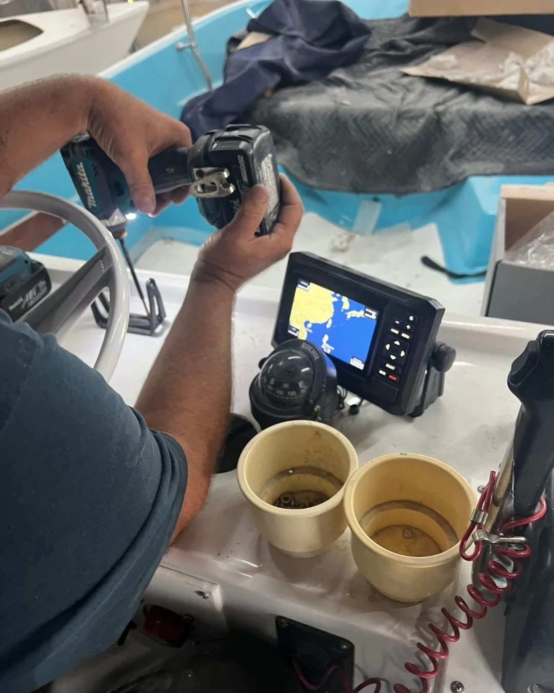 A person working on a boat, using a power drill, with a GPS navigation device and two empty coffee cups on the boat's console.