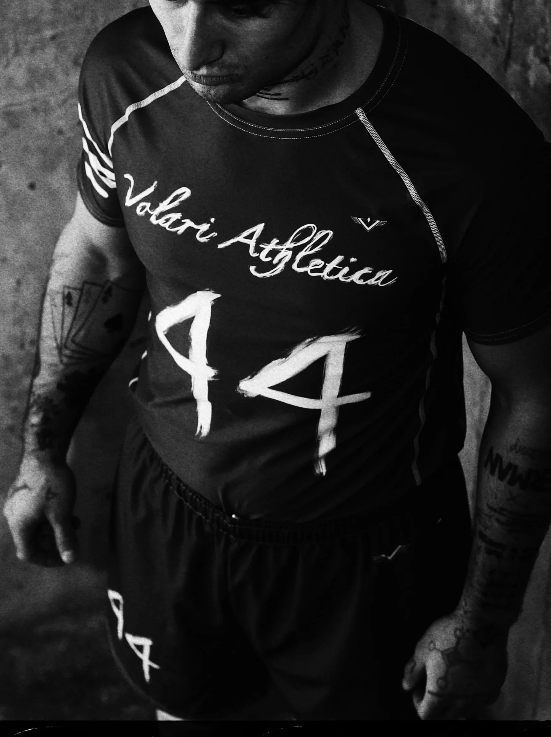 A man dressed in a BJJ Rash guard with tattoos on his arms, wearing a shirt with 'Valori Atletica' and the number 144 printed on it, standing on a rough surface.