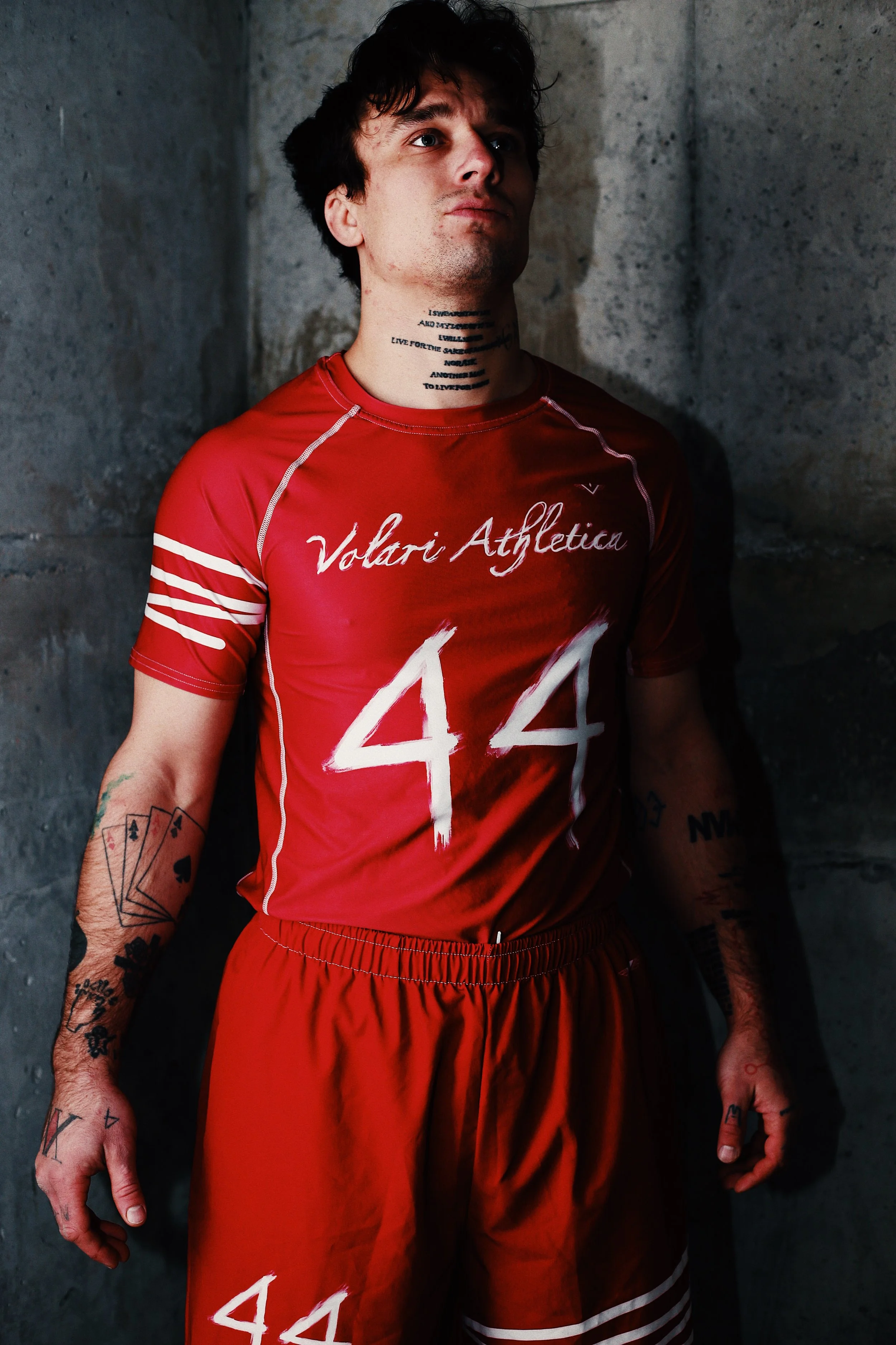 Young man with dark hair and tattoos wearing a red athletic jersey and shorts, standing against a gray concrete wall.