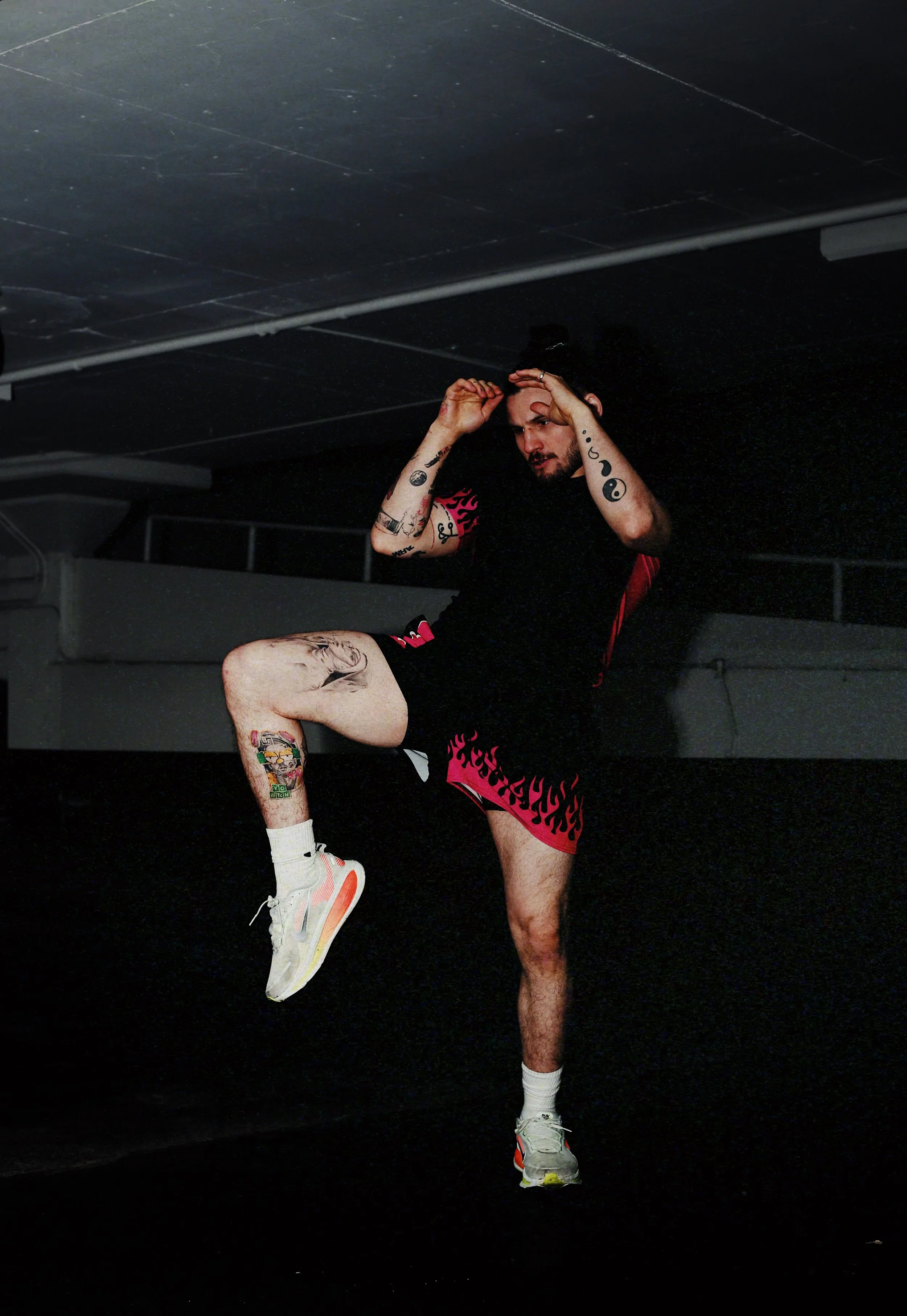 A man stretching in a dark indoor space, wearing black and red shorts, white sneakers, and with numerous tattoos on his arms and legs.
