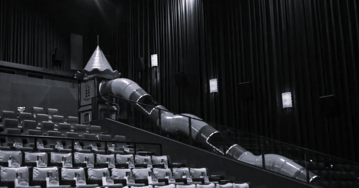 An indoor playground slide with a castle turret at the top, set inside a cinema theater with black curtains and empty seats.