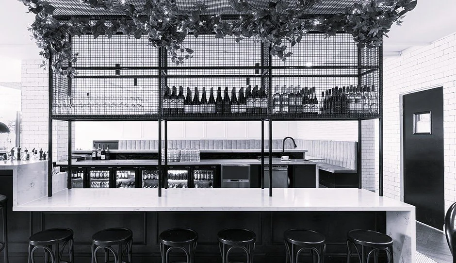 Modern bar interior with marble countertop, black stools, and metal shelving holding bottles and glassware, with a white brick wall and a black door.