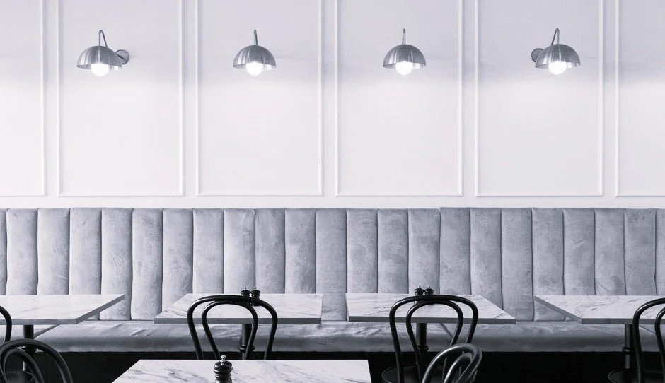 Empty modern cafe with black chairs, white marble tables, gray cushioned seating, and silver wall-mounted lamps.