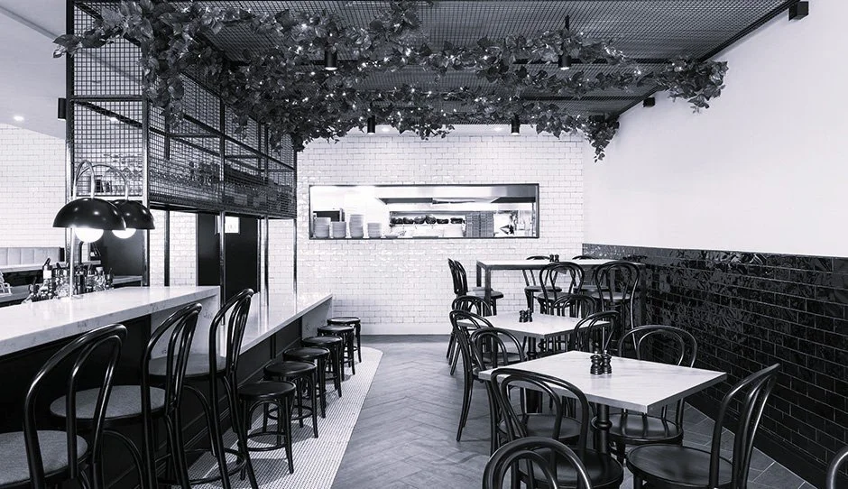 Modern cafe interior with white brick walls, black chairs, white tables, and a black and white colour scheme. Decor includes hanging foliage and a bar counter with black pendant lights.