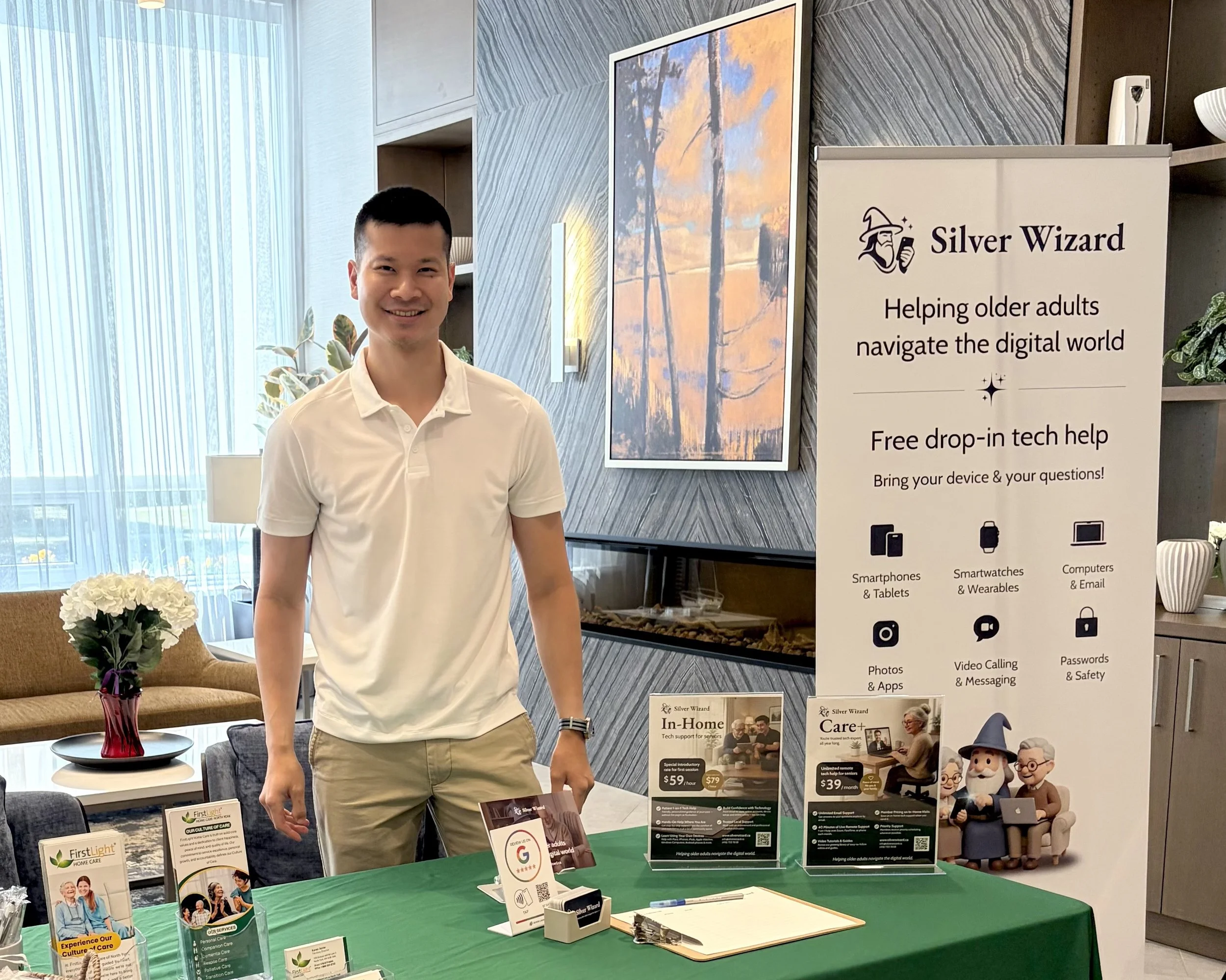 A young man in a white polo shirt standing at a booth with informational displays about digital technology support for older adults. The booth has signs and brochures, and a sign reading 'Silver Wizard.' In the background, there is a large painting of a sunset scene, a window with sheer curtains, a beige couch with a flower arrangement, and some cartoon figurines of elderly people.