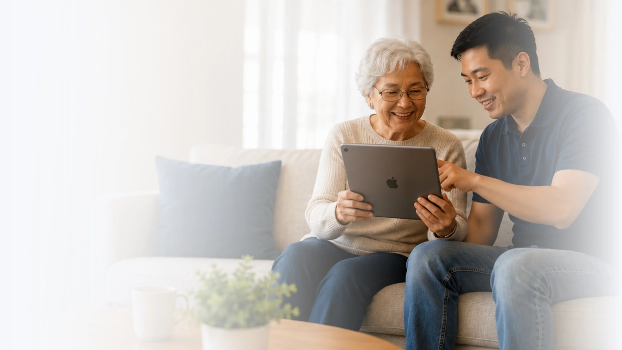 An elderly woman and a young man sitting on a couch looking at a tablet together and smiling in a bright living room.