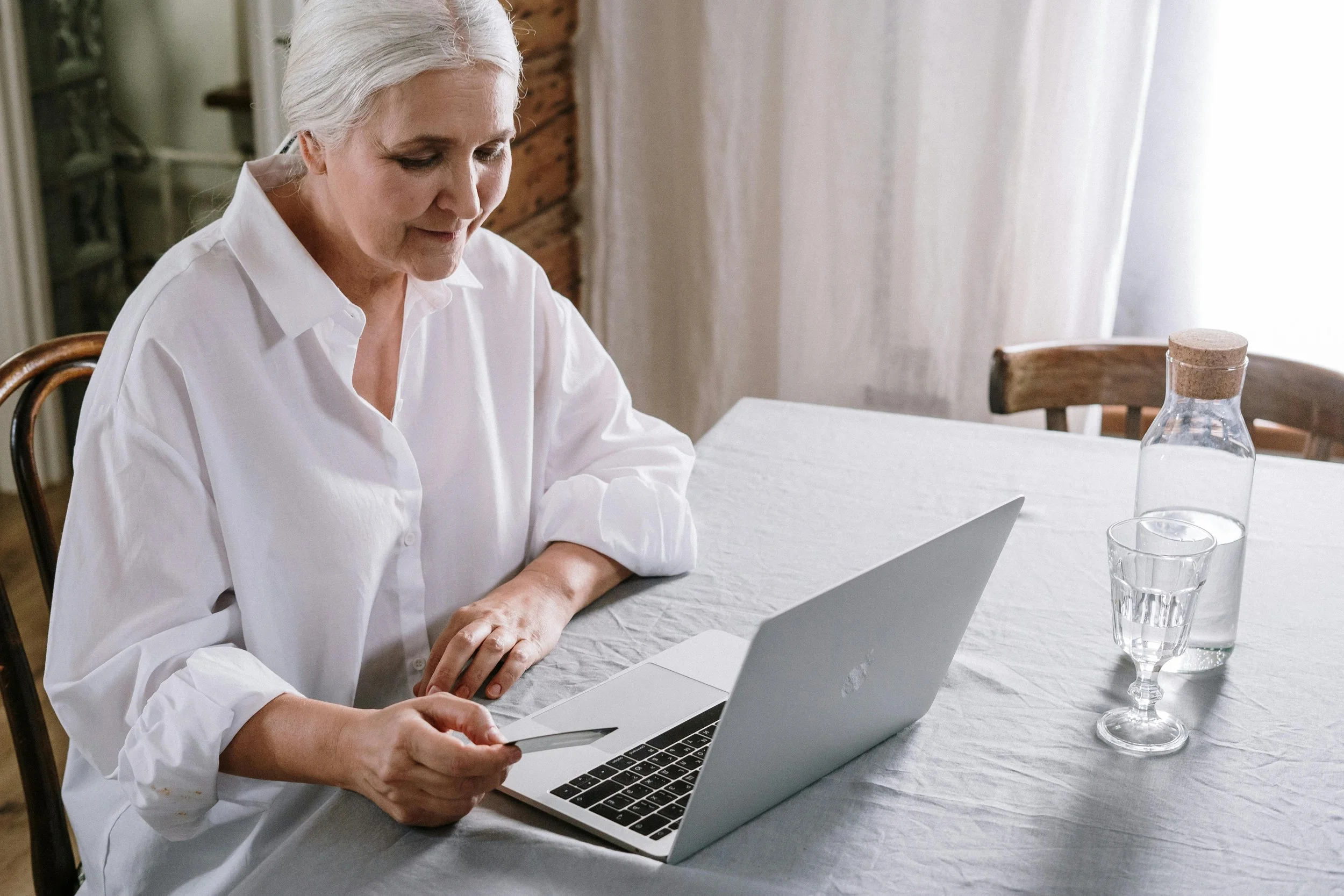 An elderly woman with gray hair wearing a white shirt sitting at a dining table, looking at her laptop, with a glass of water and a bottle on the table.
