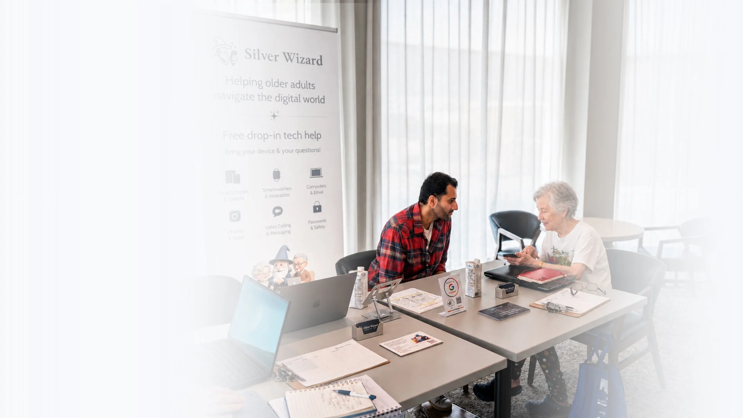 Two people sitting at a table in a bright room, looking at a smartphone together. The table has laptops, notebooks, and drinks. A banner in the background reads "Silver Wizard" and mentions helping older adults with digital technology.