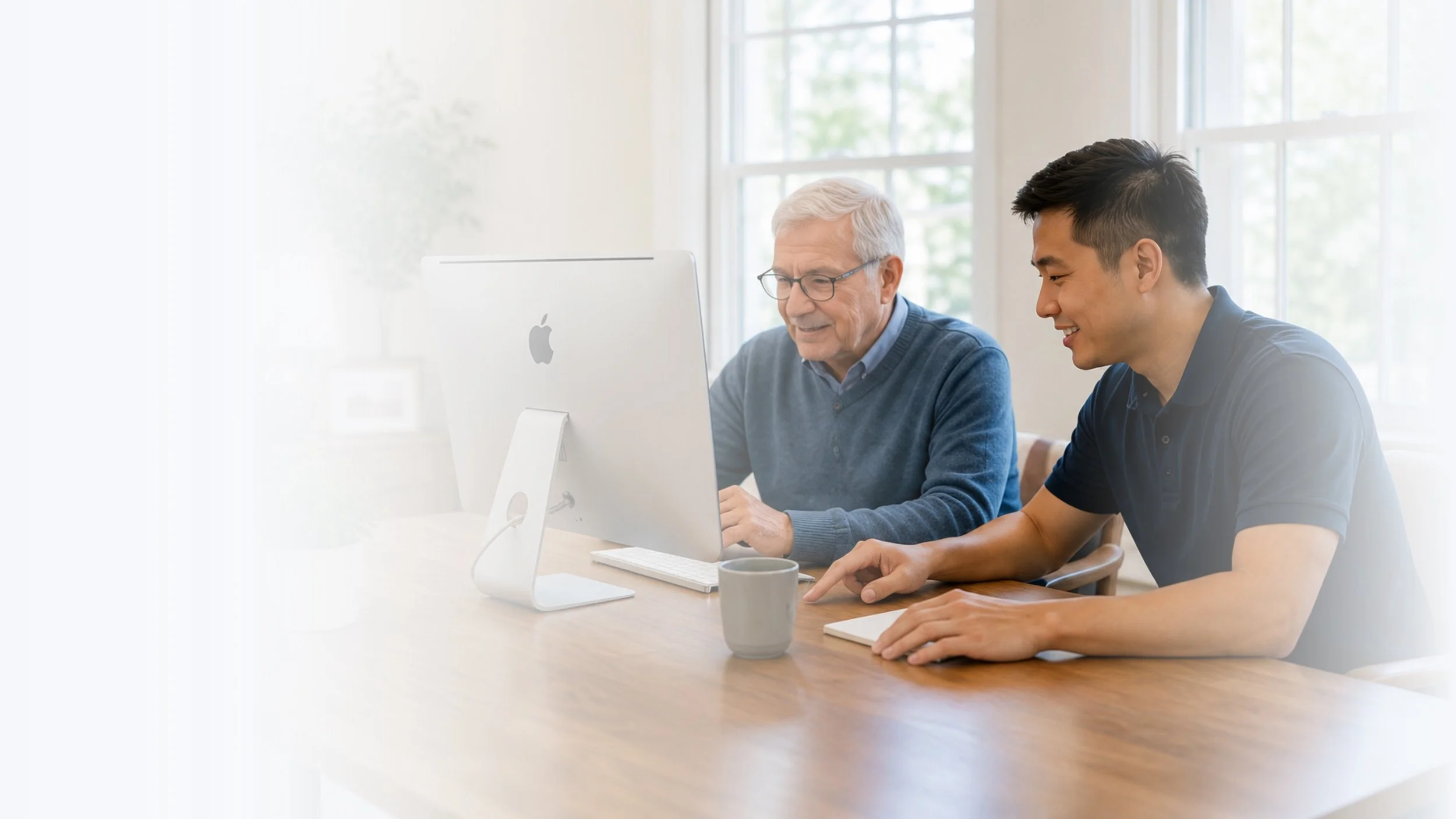 Two men sitting at a wooden table looking at a computer screen, in a bright room with large windows, one older man with glasses and gray hair, the other younger man with dark hair, both smiling.