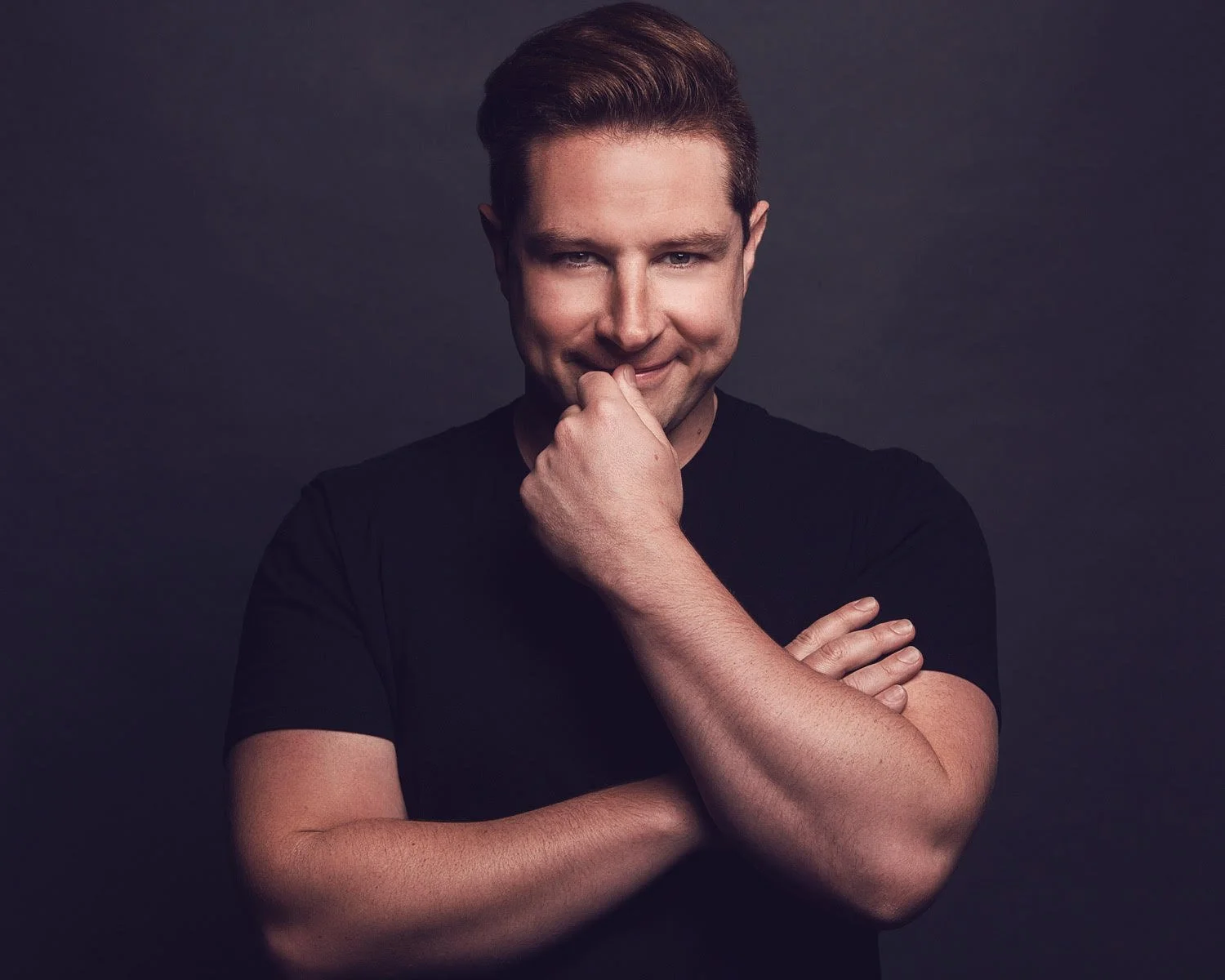 Portrait of a man with red hair wearing a black t-shirt, smiling and posing with one arm crossed and the other hand near his face against a dark background.