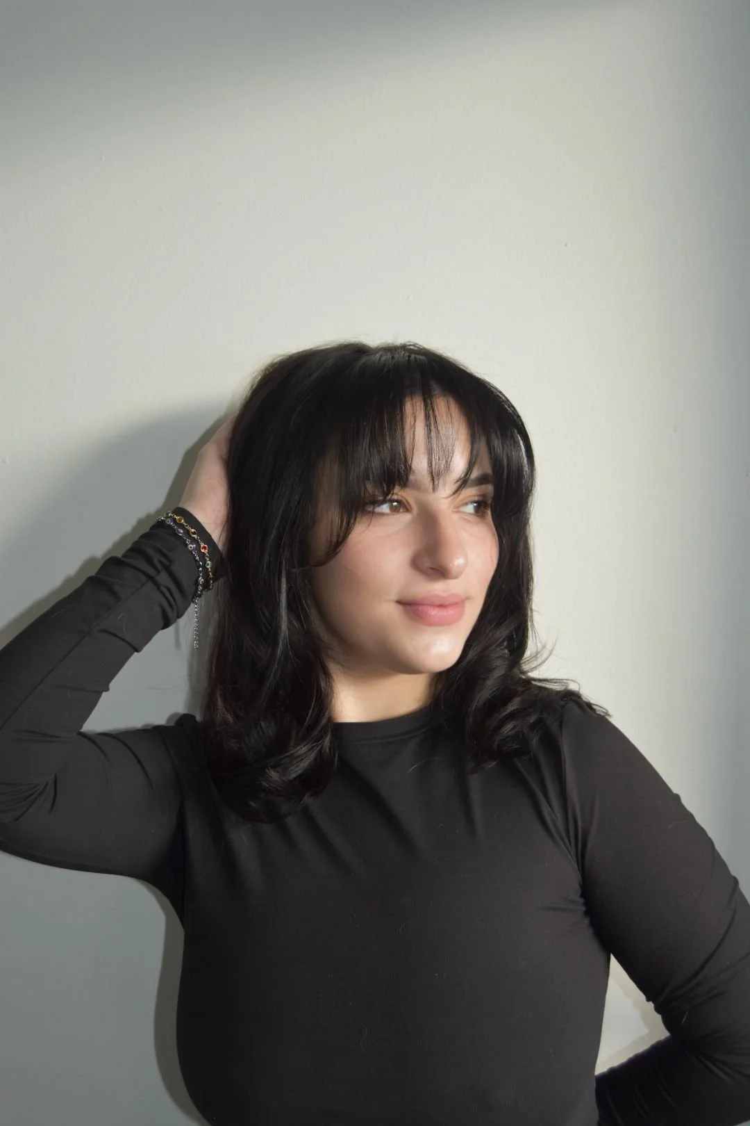 A woman with shoulder length dark hair and bangs, wearing a black long-sleeve top, standing against a plain wall, looking slightly to her right with a gentle smile.