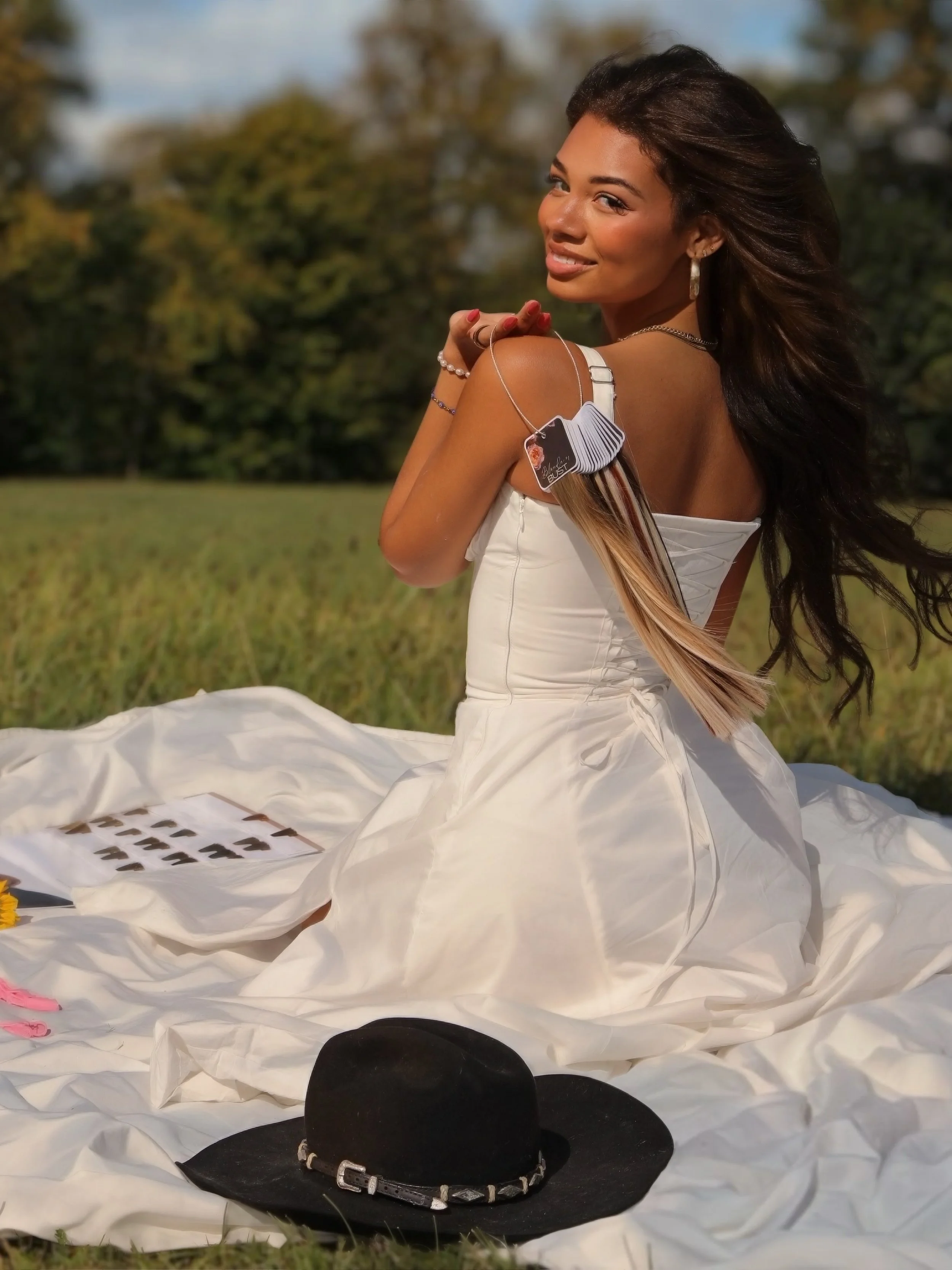 A woman in a white dress with long hair sitting on a white blanket outdoors, smiling, with a black hat and photos of hair color swatches nearby.