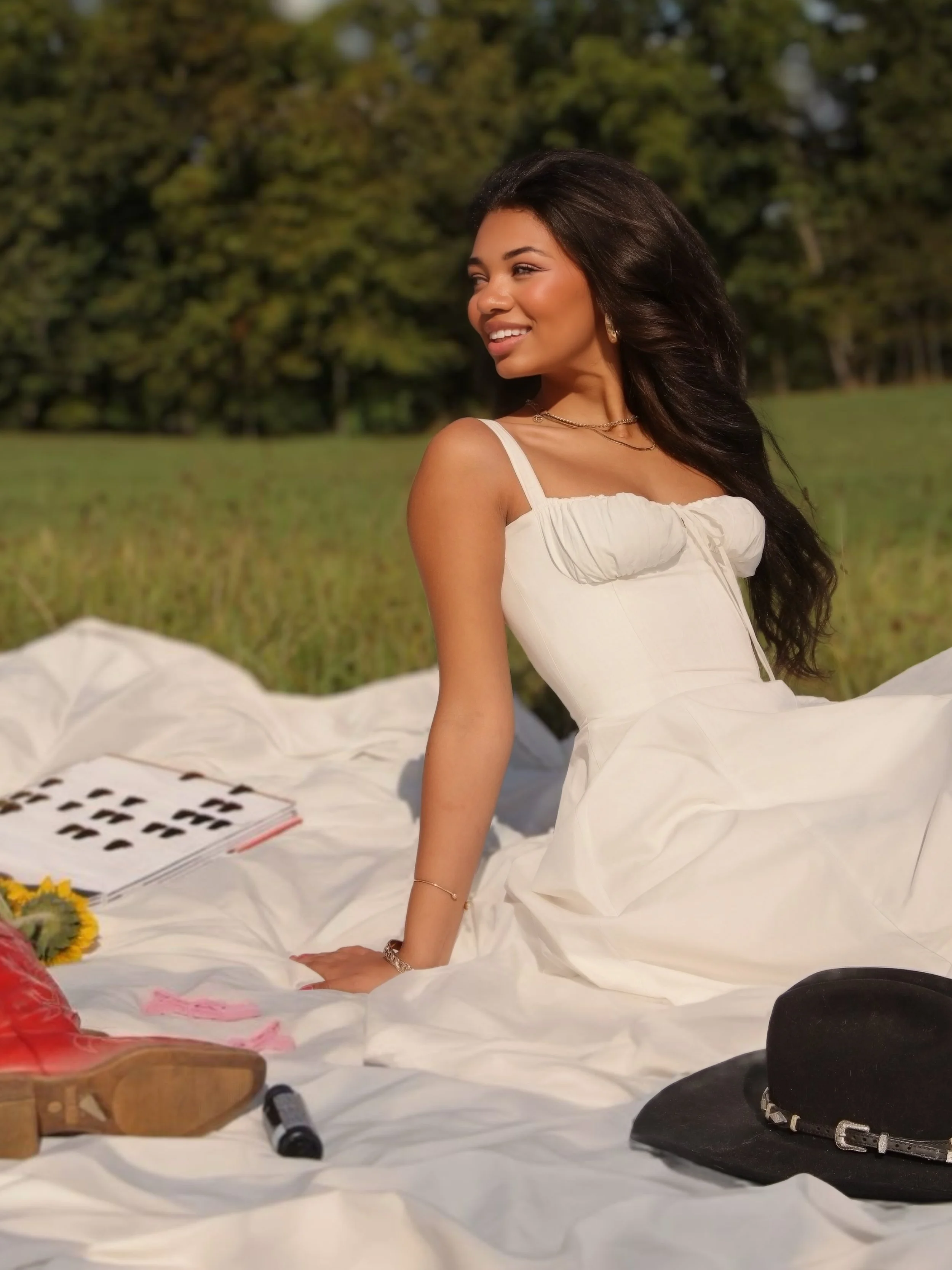 A woman sitting on a white blanket outdoors in a grassy field with trees in the background, smiling and looking to the side. There are items on the blanket, including a black hat, a book, a lipstick, and a pair of cowboy boots.
