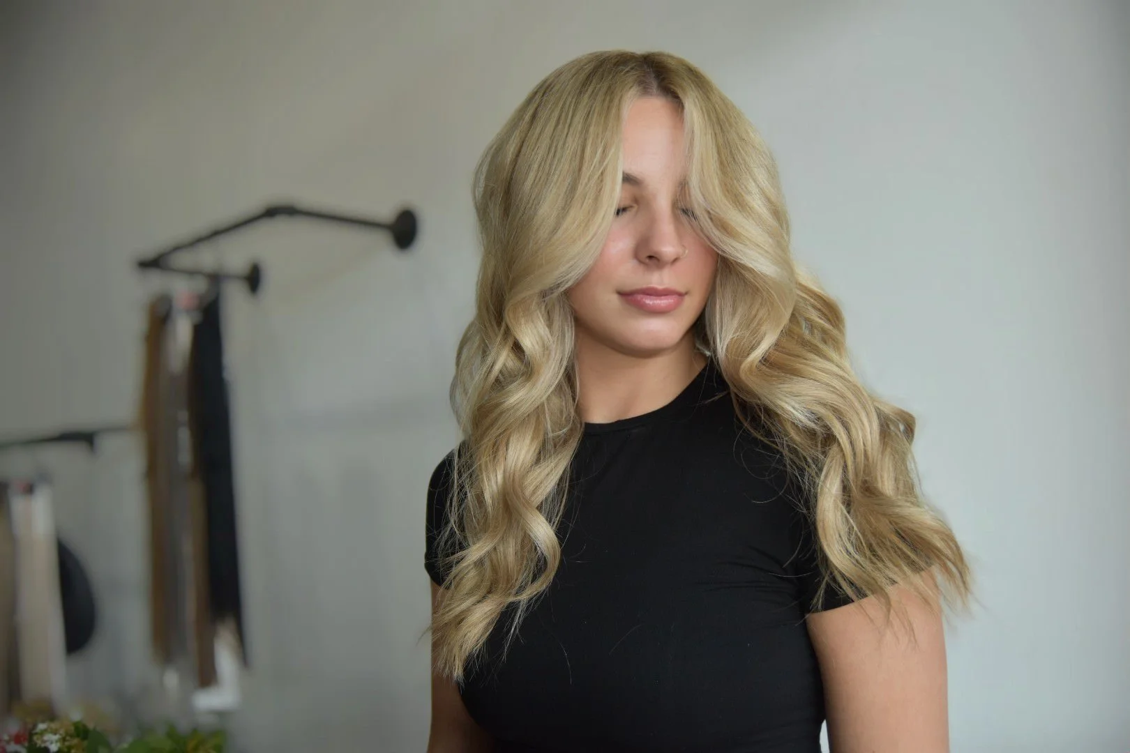 A young woman with long, wavy blonde hair wearing a black t-shirt standing indoors with a neutral expression and eyes closed. There is a blurred background with some equipment hanging on a wall.