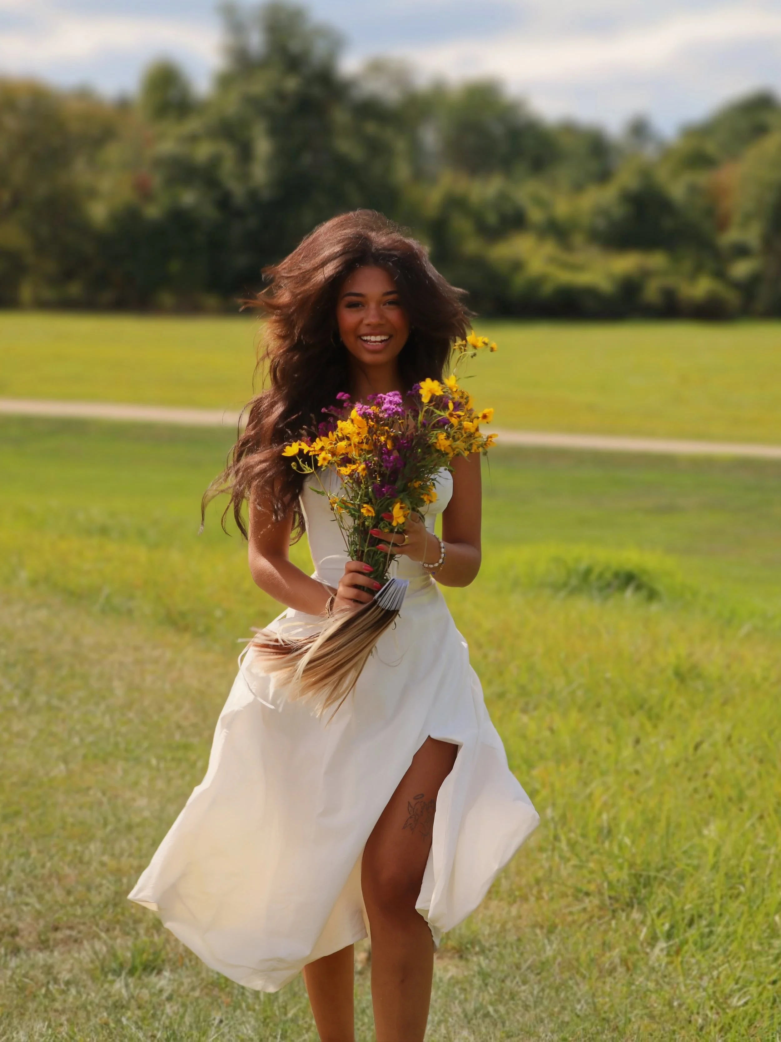 A woman smiling while running through a grassy field, holding a colorful bouquet of flowers, wearing a white dress with a high slit revealing a tattoo on her thigh.