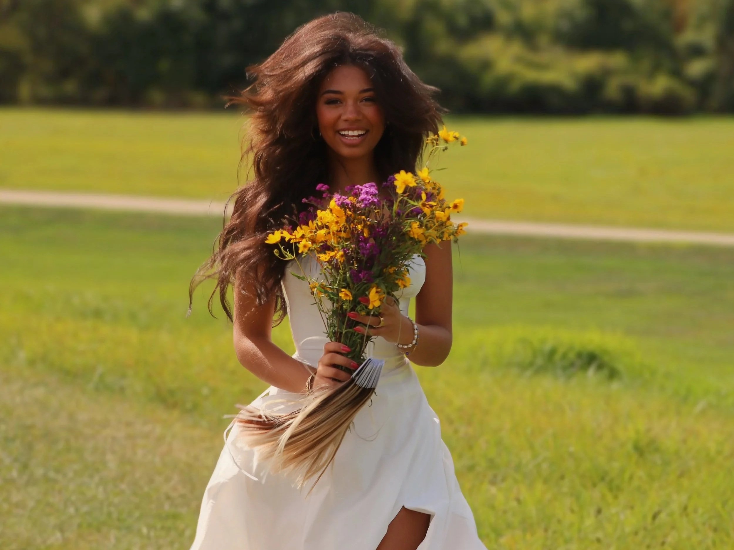 Smiling woman in a white dress holding a colorful bouquet of flowers in an outdoor grassy area.