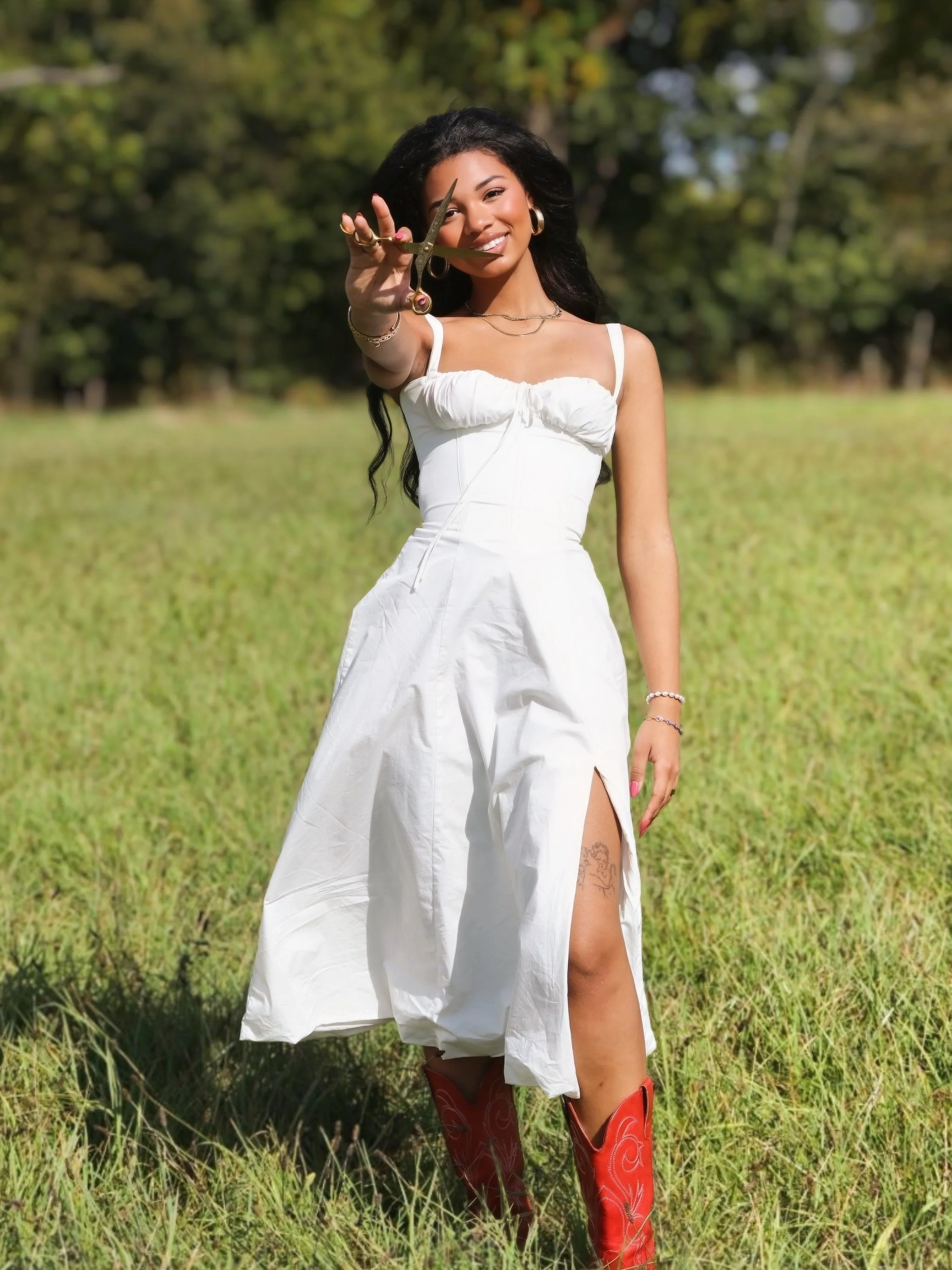 A woman in a white dress, with black hair, is standing outdoors in a grassy field with trees in the background. She is smiling and holding a pair of scissors toward the camera, wearing red boots and jewelry.