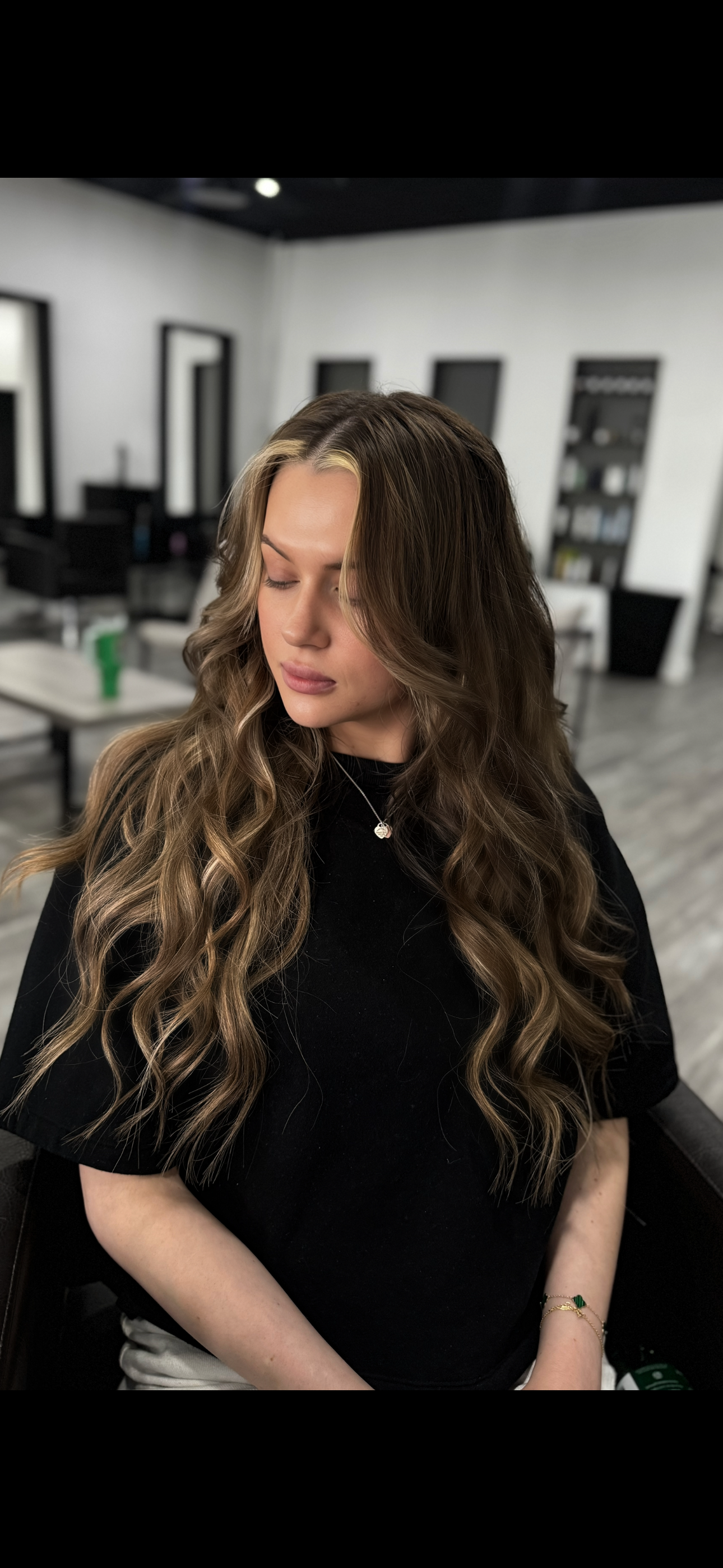 Woman with long, wavy brown hair sitting in a salon chair with her eyes closed, in a modern salon with black and white decor.