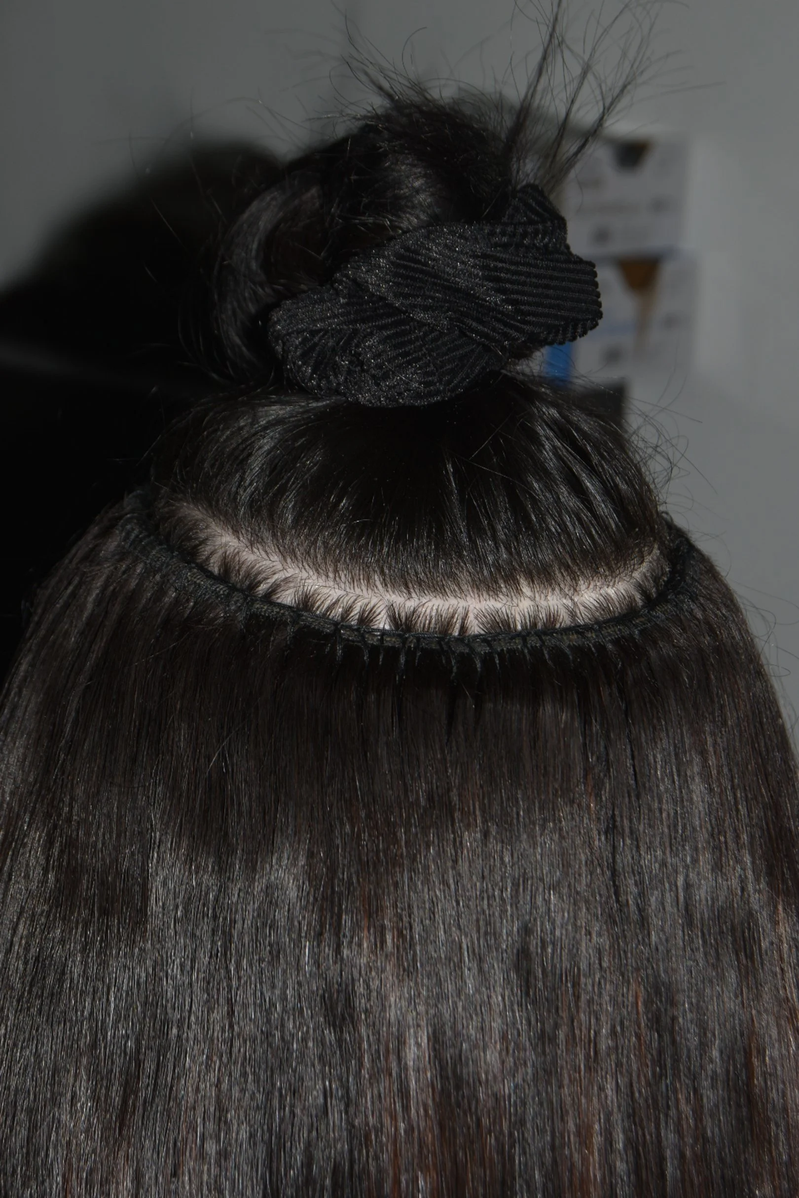 Close-up of a woman with straight dark hair, partially sewn in a hair extension process, with a hair artist securing extensions with a black scrunchie.