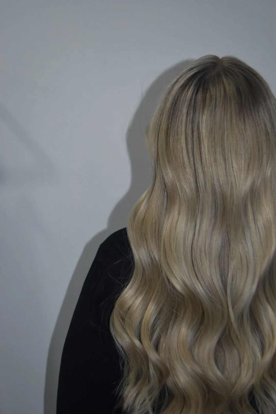 A person with long, wavy blonde hair standing against a plain wall, casting a shadow.