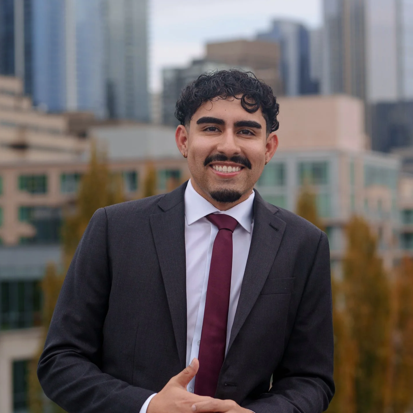 A smiling man in a dark suit with a white shirt and burgundy tie standing outdoors in front of a cityscape with tall buildings.