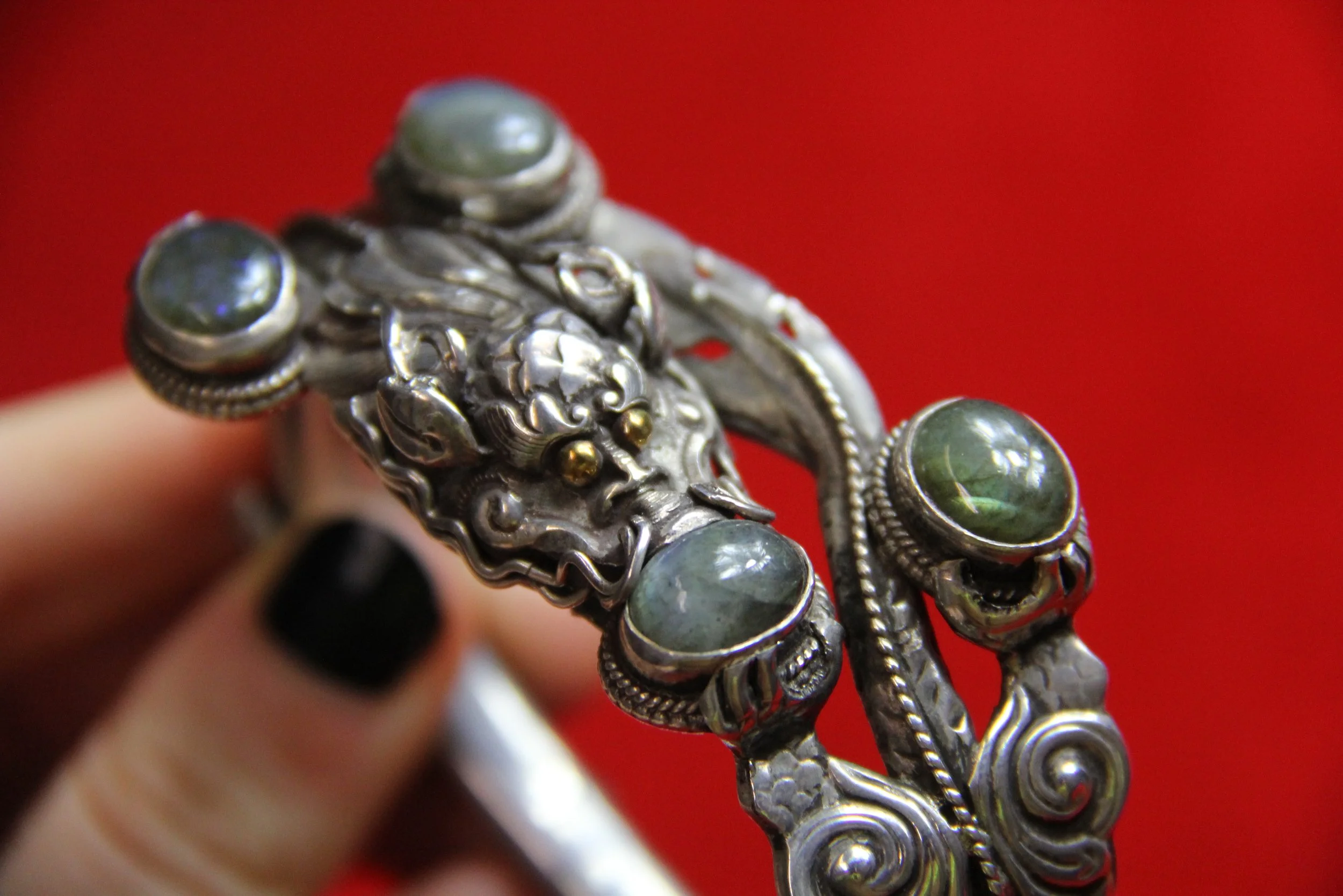 Close-up of an ornate silver bracelet with a lion's head motif and green stones, held by a hand with black nail polish against a red background.