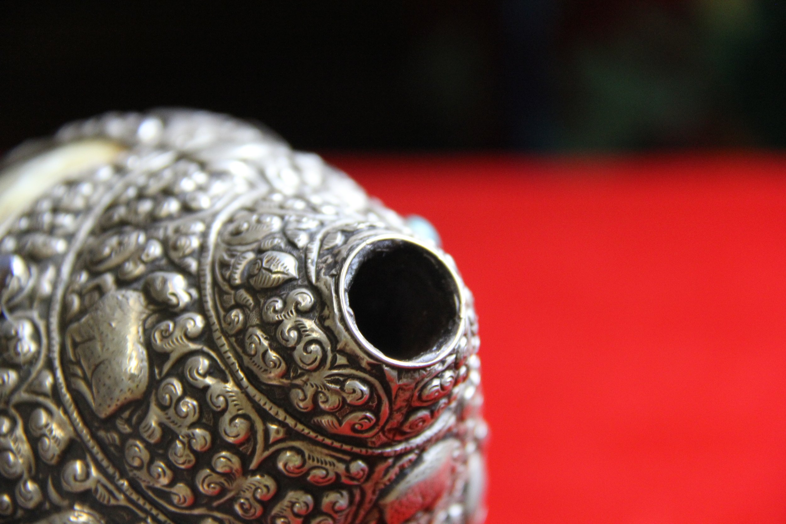 Close-up of an ornate, silver decorative object with intricate patterns, and a hollow opening, set against a background with black and red colors.