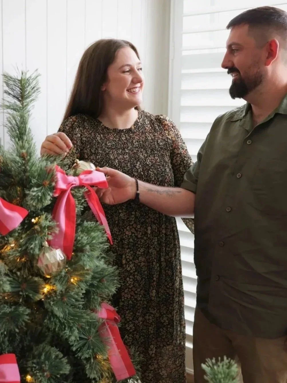 A couple is exchanging a gift near a decorated Christmas tree with pink bows and ornaments, indoors by a window with white blinds, smiling at each other.