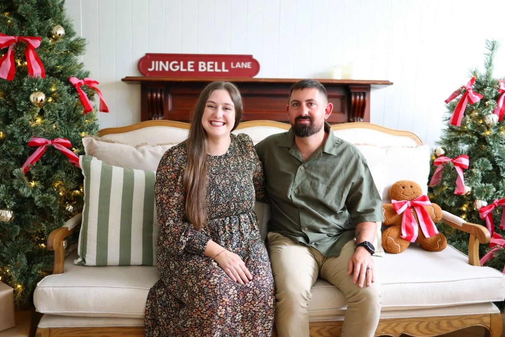 A smiling woman and a man sitting on a beige sofa, surrounded by Christmas trees decorated with red ribbons and golden ornaments, in a festive Christmas setting. Christmas Mini Session.