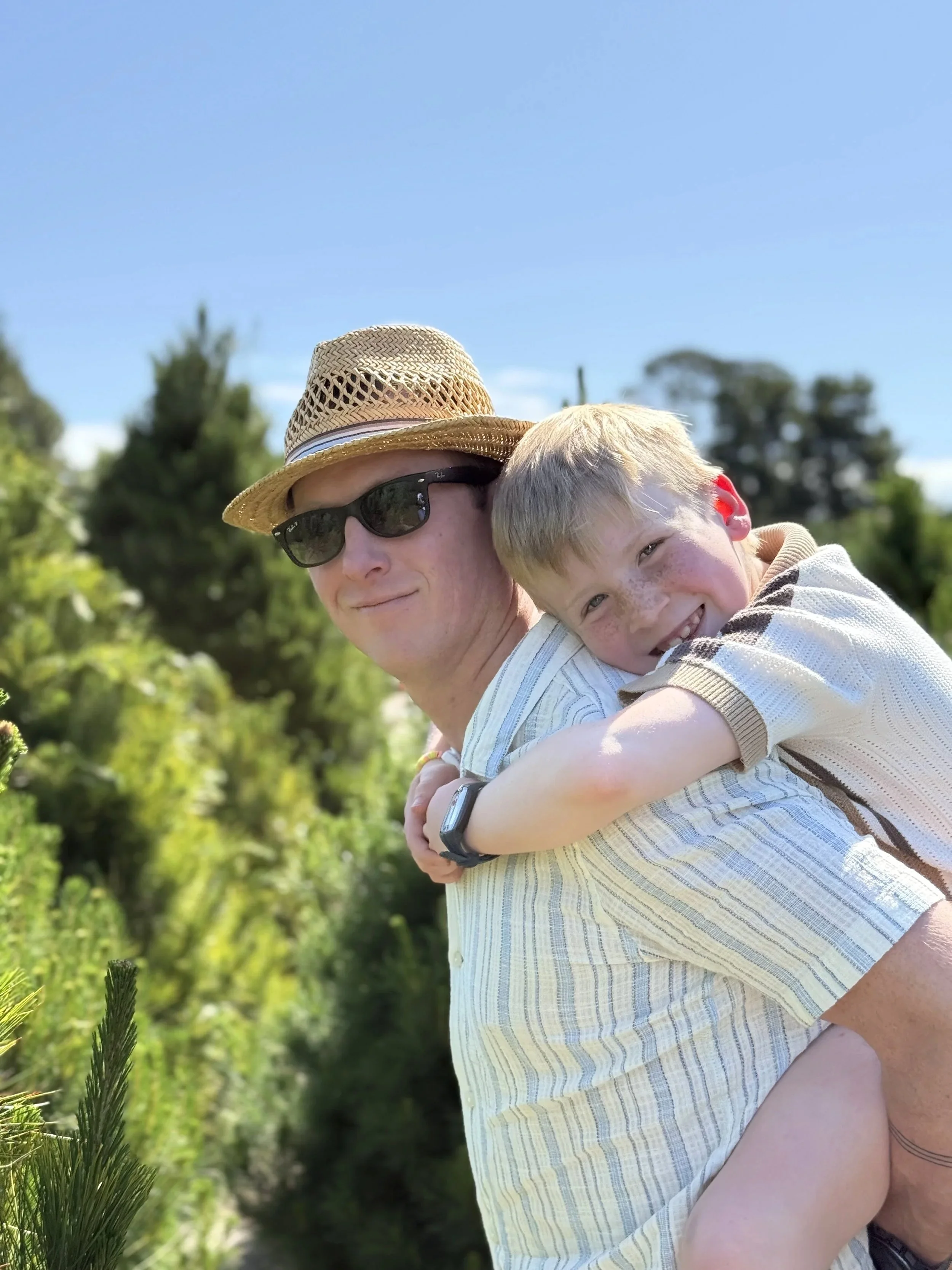 A man wearing sunglasses and a straw hat carrying a young boy on his back outdoors. The boy is smiling and hugging the man, both surrounded by green trees under a clear blue sky.