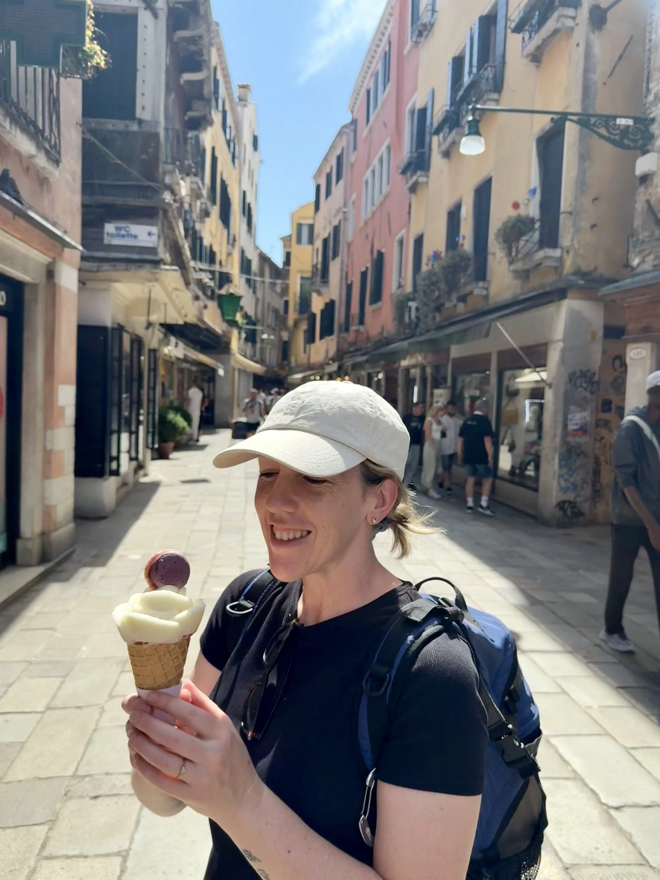 A woman smiling and holding an ice cream cone on a sunny street with colorful buildings and pedestrians.
