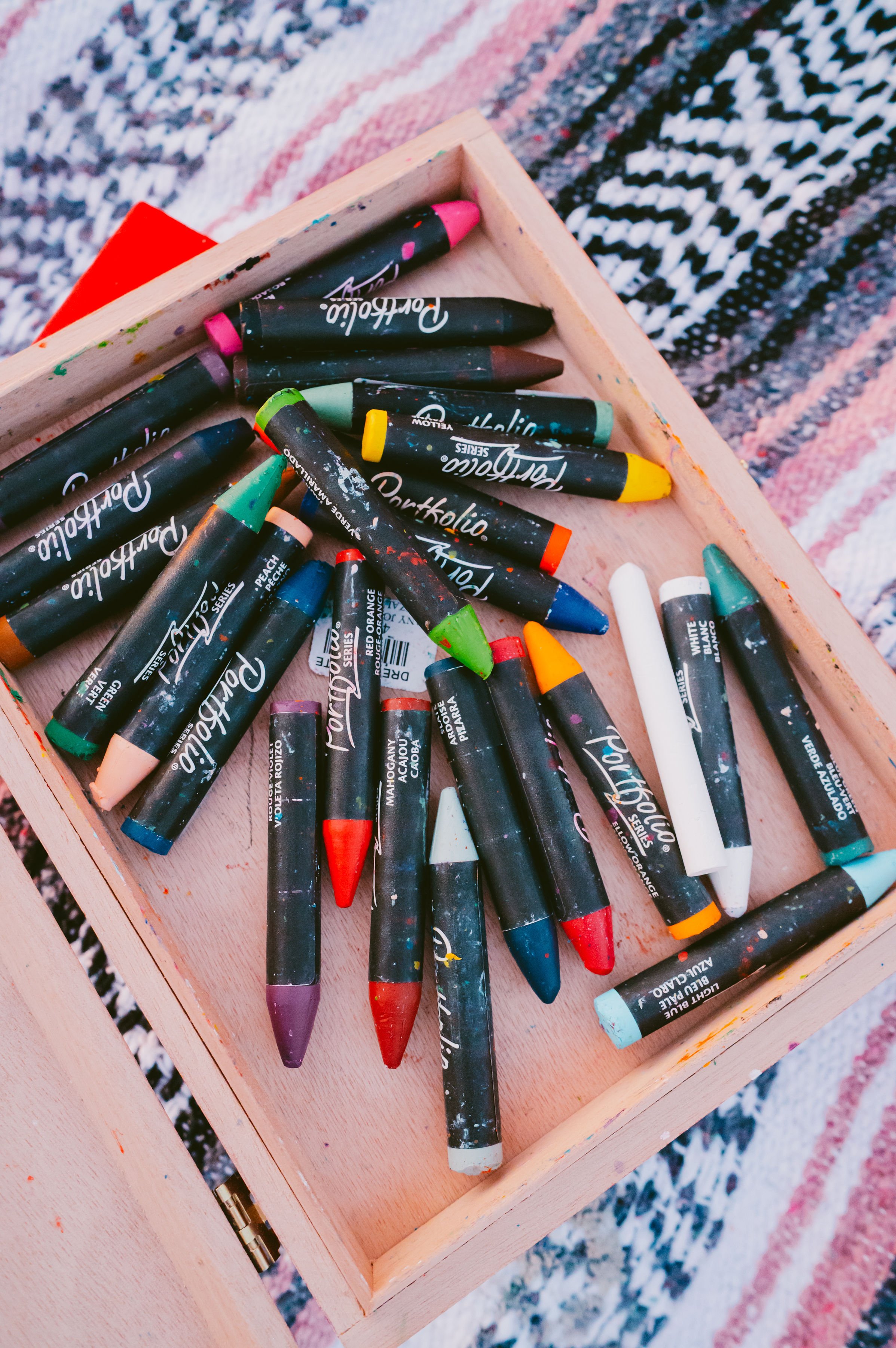 A wooden tray containing various colorful crayons with a patterned textile in the background.
