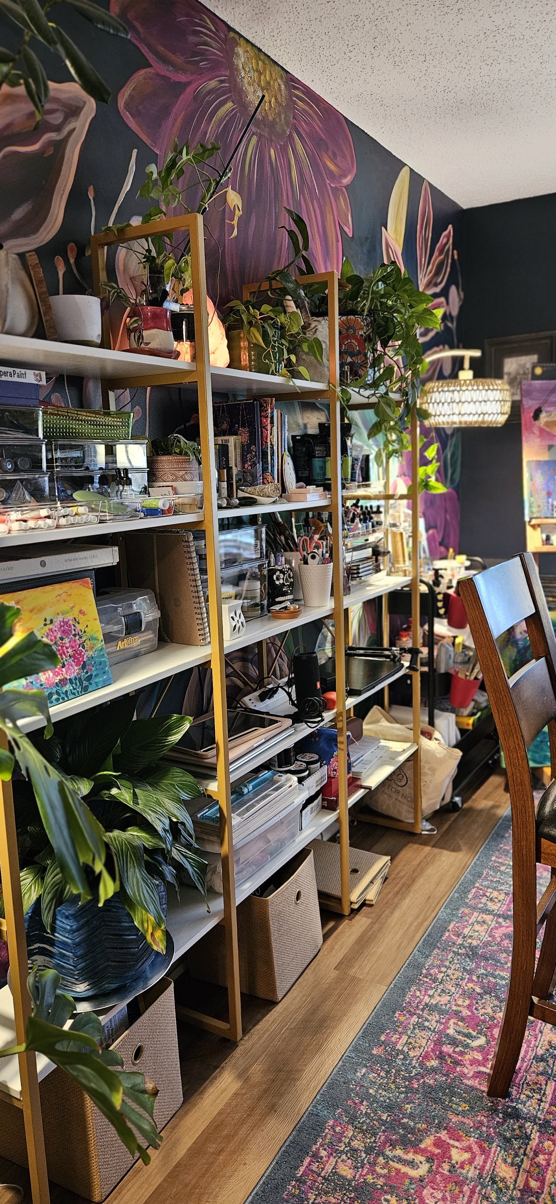 A colorful art studio with black walls featuring a large pink and purple floral mural, filled with shelves of art supplies, plants, and decorative items, next to a patterned area rug and wooden chair.