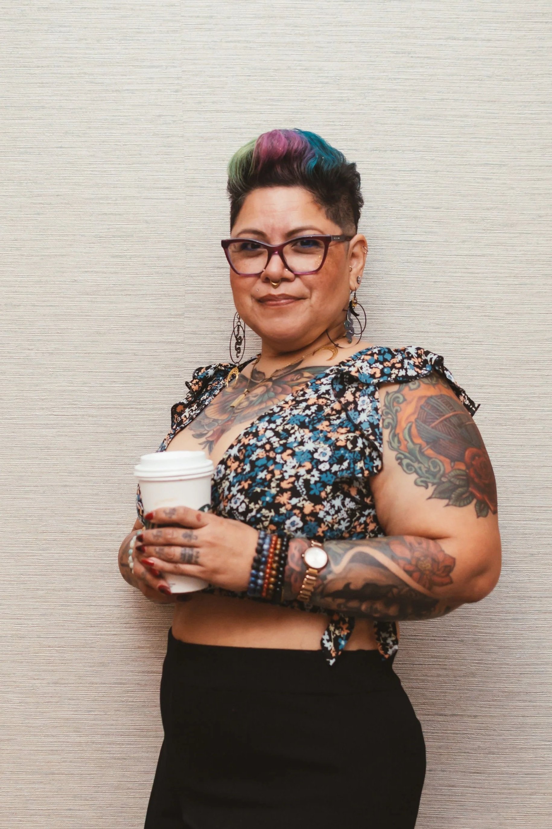A woman with short, colorful hair, glasses, and tattoos holding a coffee cup, standing against a beige textured wall.