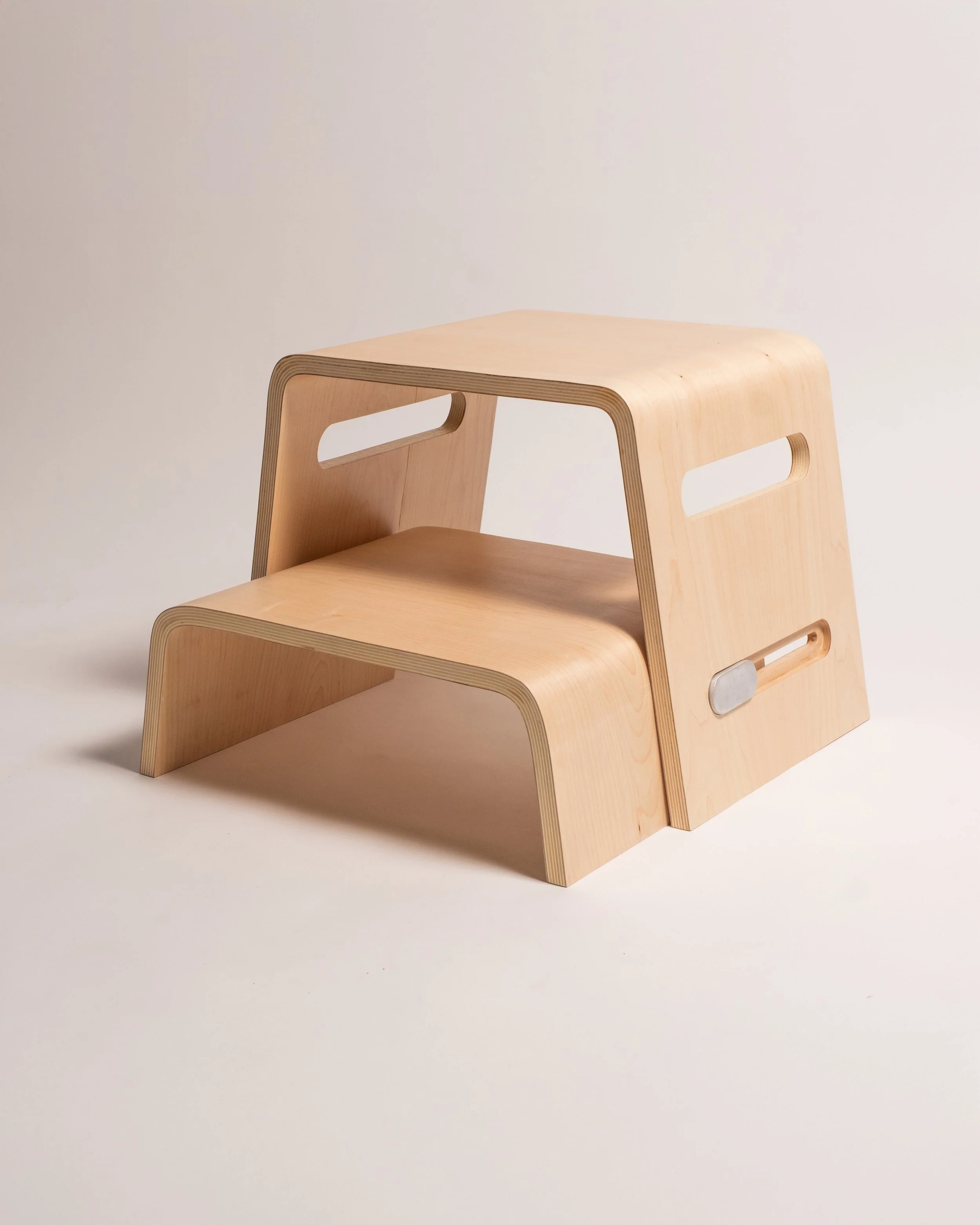 Two minimalist wooden stools stacked together with cutout handles, on a plain light background.