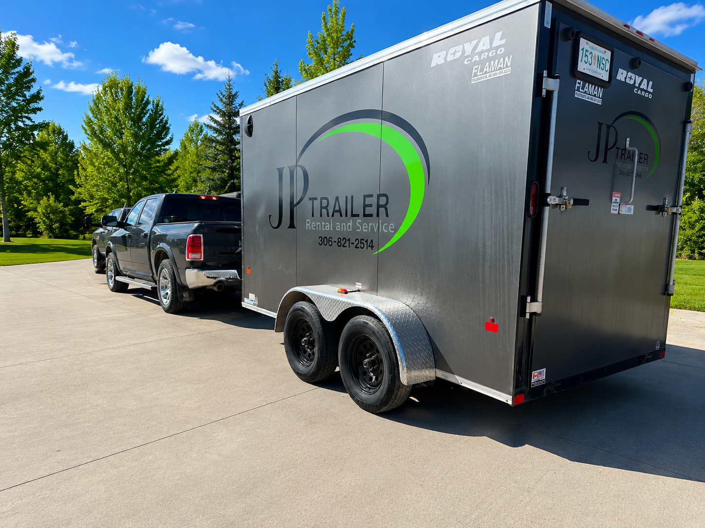 A black pickup truck attached to a gray enclosed trailer from JP Trailer Rental and Service parked on a concrete driveway with green trees in the background and blue sky above.