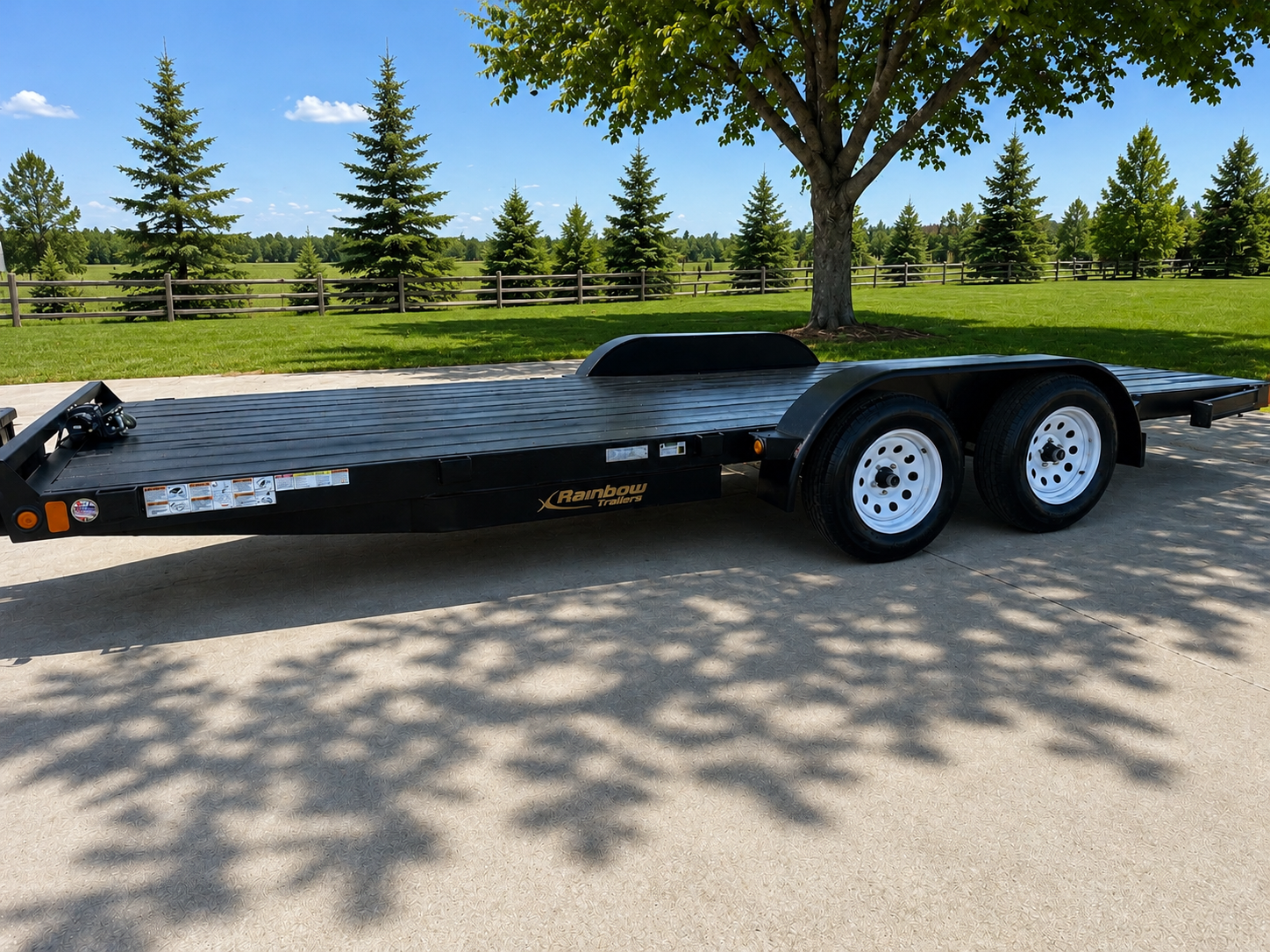 A black Rainbow Trailer flatbed trailer parked on a concrete surface under a tree casting shadow, with a park-like background of green grass, trees, and a clear blue sky.