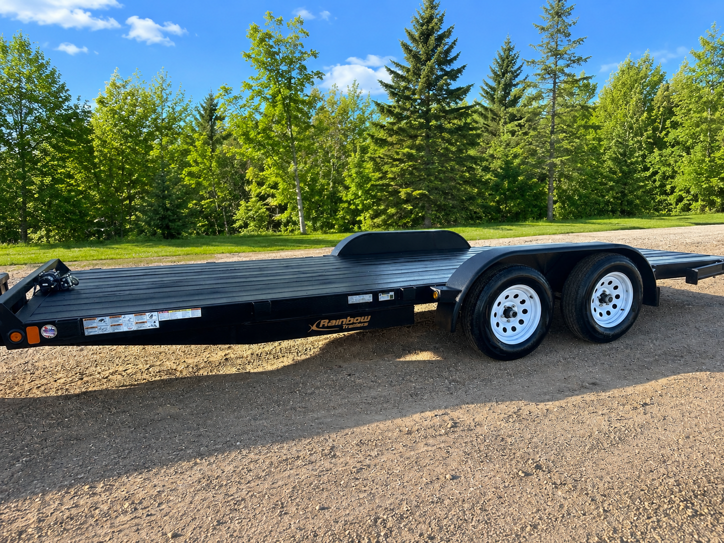 Black flatbed trailer with dual axles parked on dirt ground, with green trees and blue sky in the background.