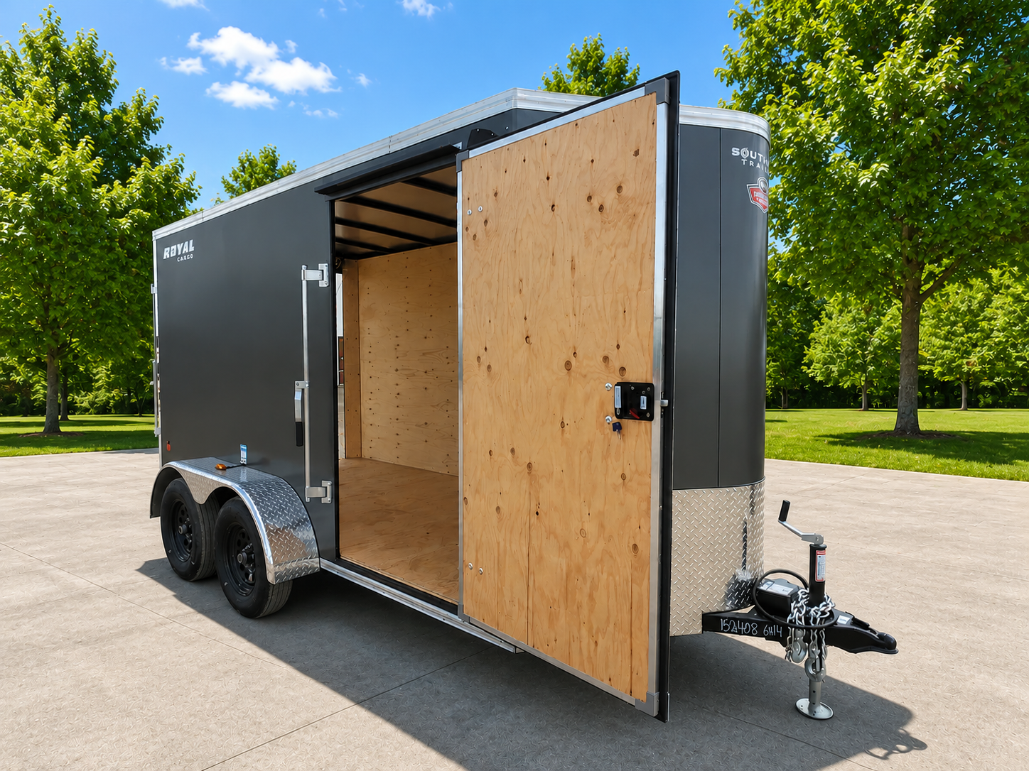 Black enclosed trailer with open side door, made of wood interior, parked on concrete, surrounded by green trees and blue sky.