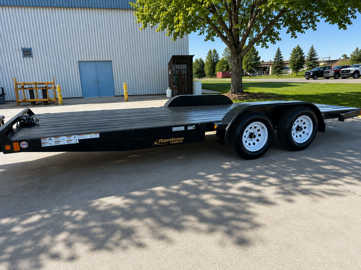 Black flatbed trailer with two axles parked on a concrete surface under a tree, with a metal building, green grass, and trees in the background.