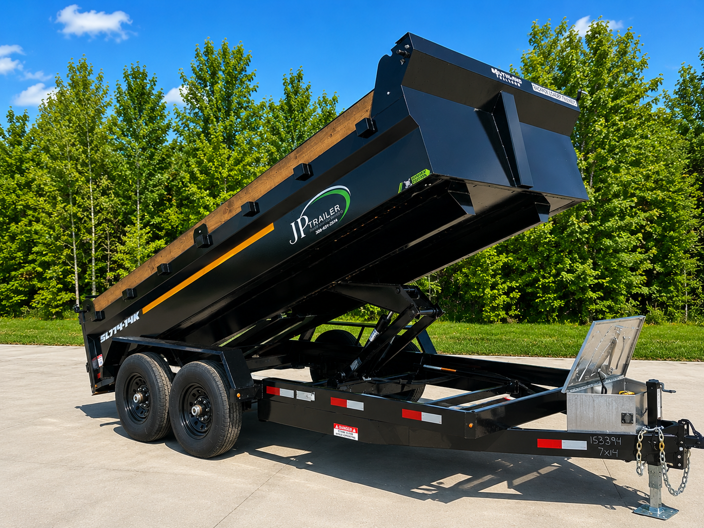 Black dump trailer with hydraulic lift raised, parked on concrete surface with green trees and blue sky in the background.