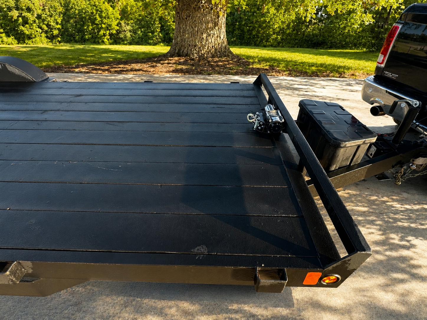 A black flatbed trailer with a wood deck and a hitch, parked outdoors near a tree and a grassy area. A black toolbox is mounted on the front of the trailer.