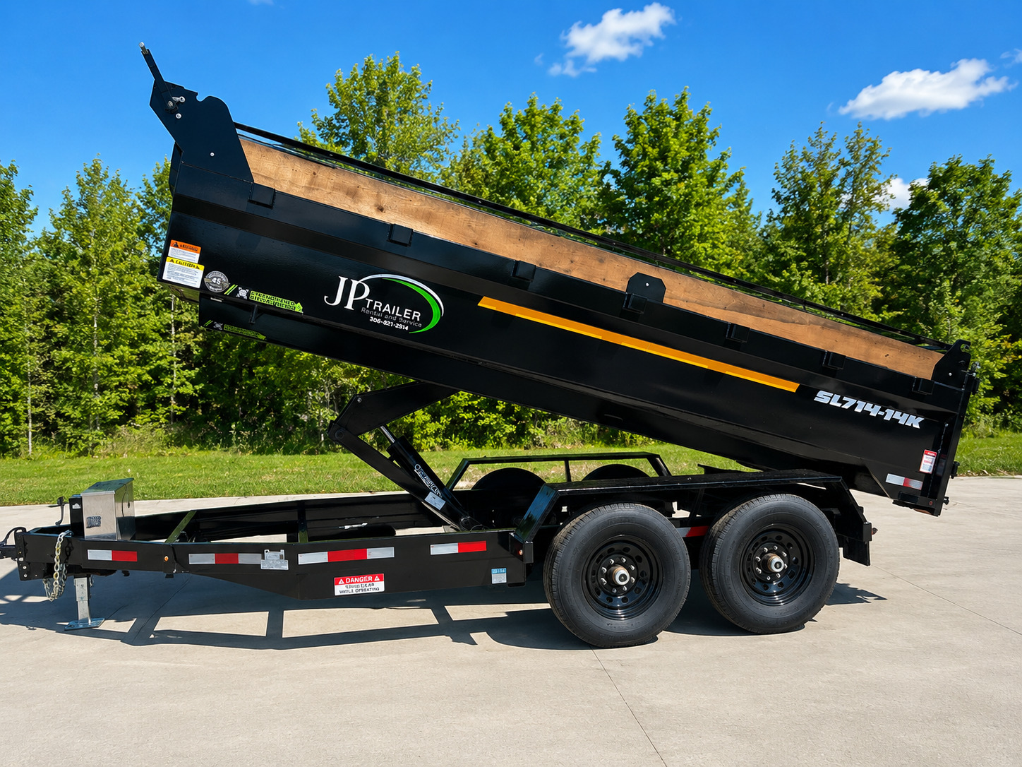A black dump trailer with a wooden bed, raised and tilted for unloading, on a concrete surface with trees and blue sky in the background.