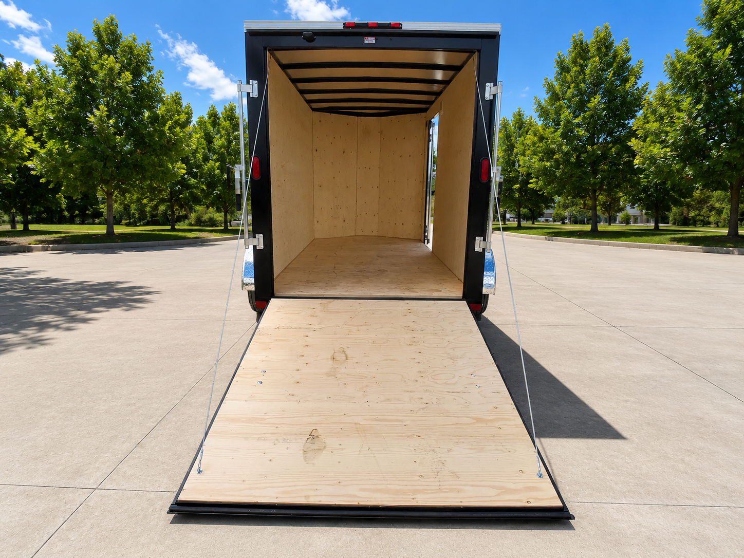 Empty black enclosed trailer with a lowered wooden ramp door, parked on a driveway with trees and a blue sky in the background.