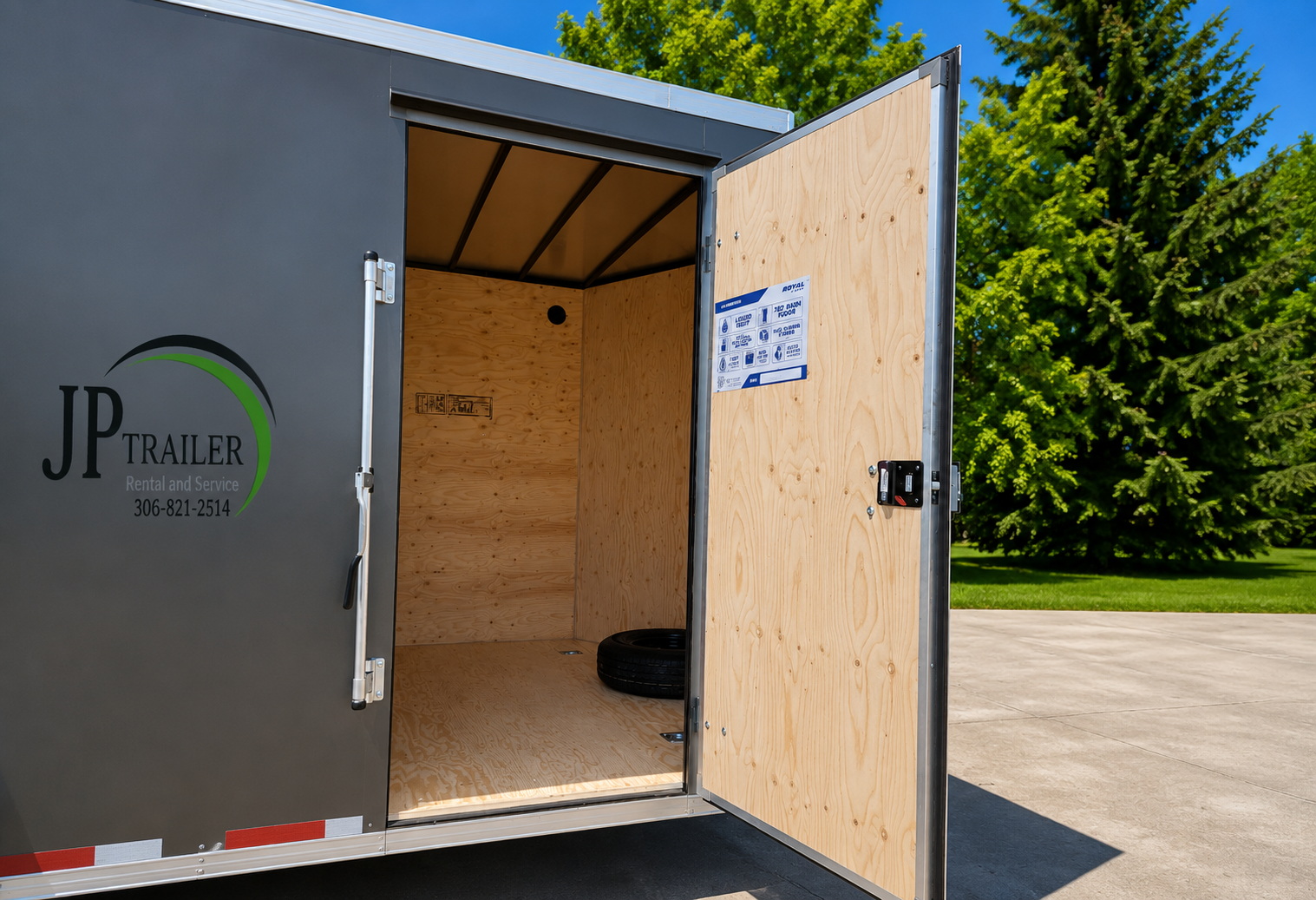 Open cargo trailer with wooden interior, black tire inside, metal locking mechanisms, and a label on the door, parked on a driveway with trees and a blue sky in the background.