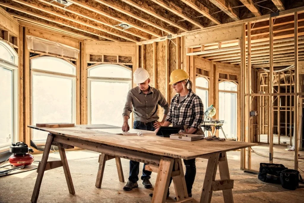 Two construction workers, a man and a woman, wearing safety helmets, looking at blueprints on a large table in a partially built house with exposed wooden framing and large windows.