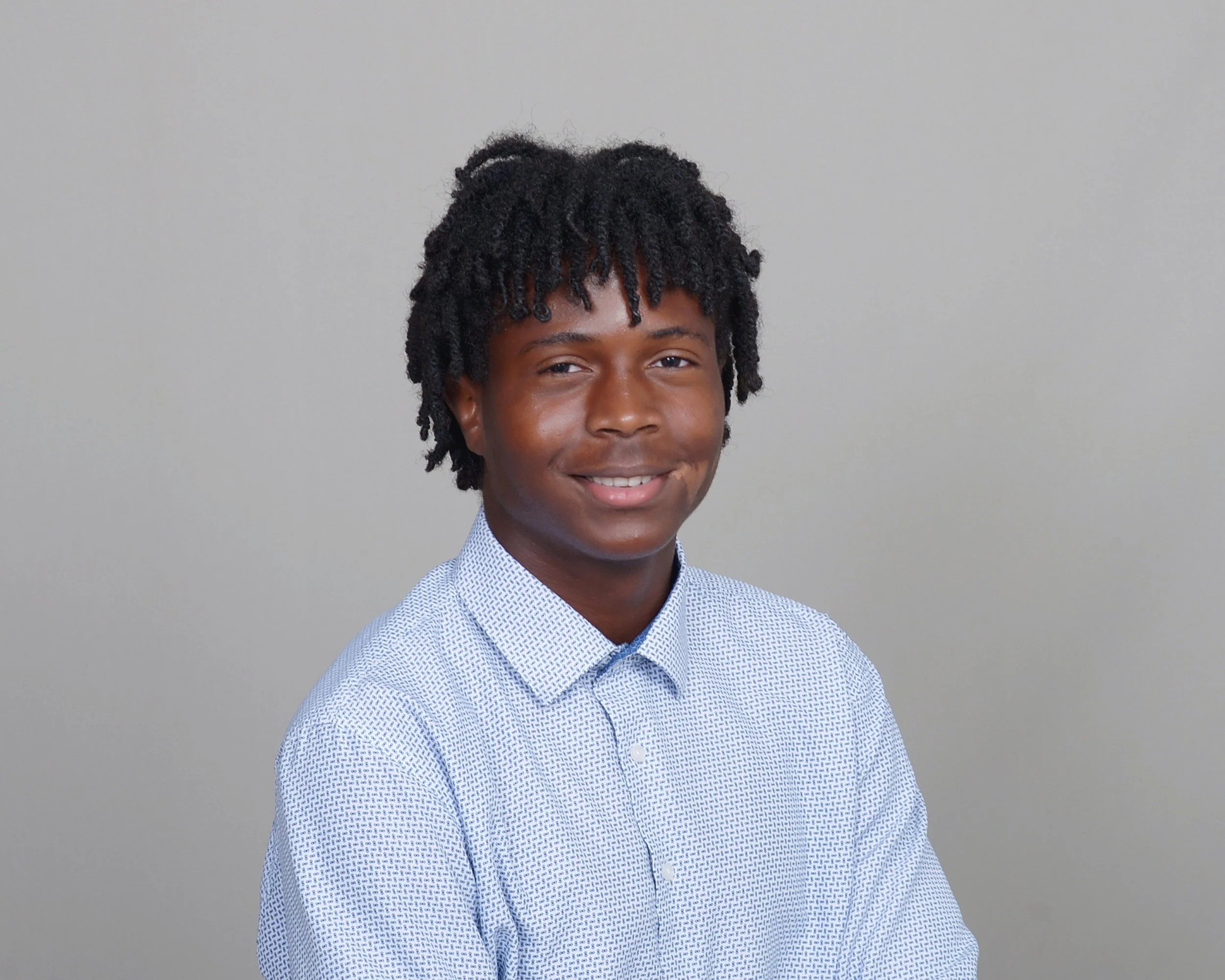 Young man with dark skin, curly hair, wearing a light blue patterned dress shirt, smiling against a plain gray background.