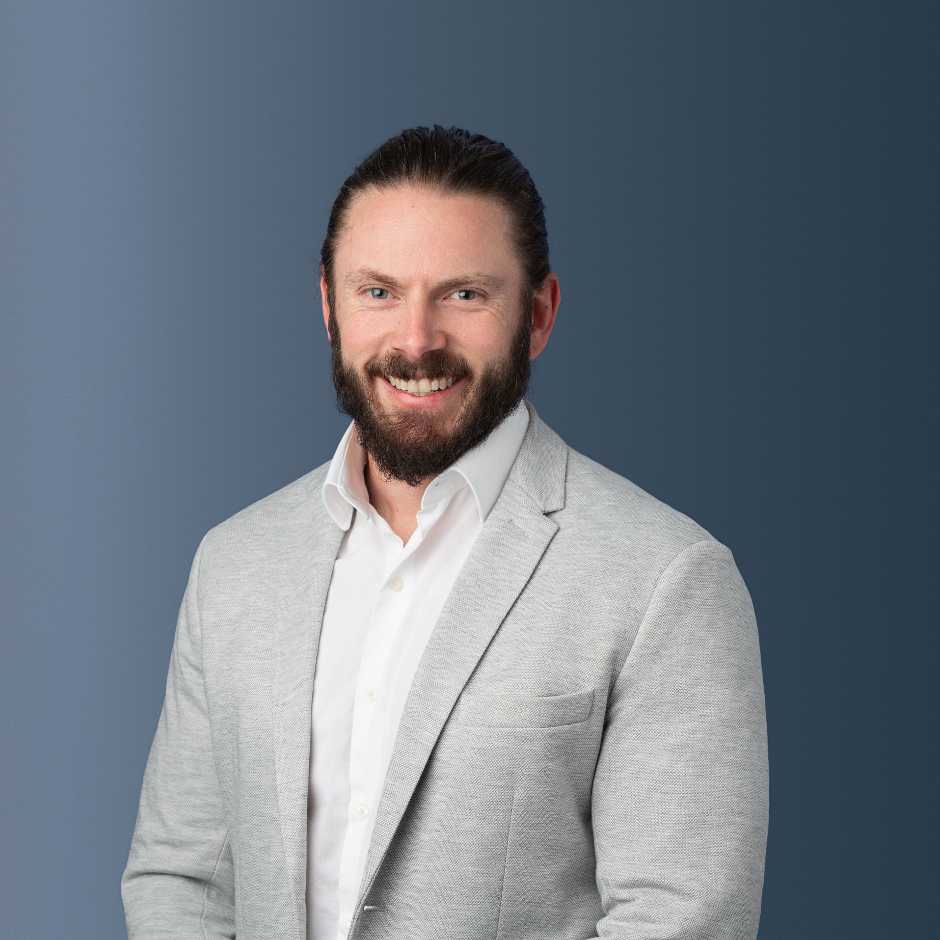 Portrait of a smiling man with a beard, wearing a light gray blazer and white shirt, against a blue gradient background.